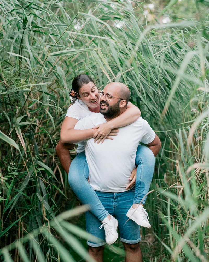 Homme portant sa compagne sur son dos en riant au milieu de la végétation aux Jardins de l'Argentor à Nanteuil-en-Vallée lors d'une séance photo de couple par Alexandre Topolewski