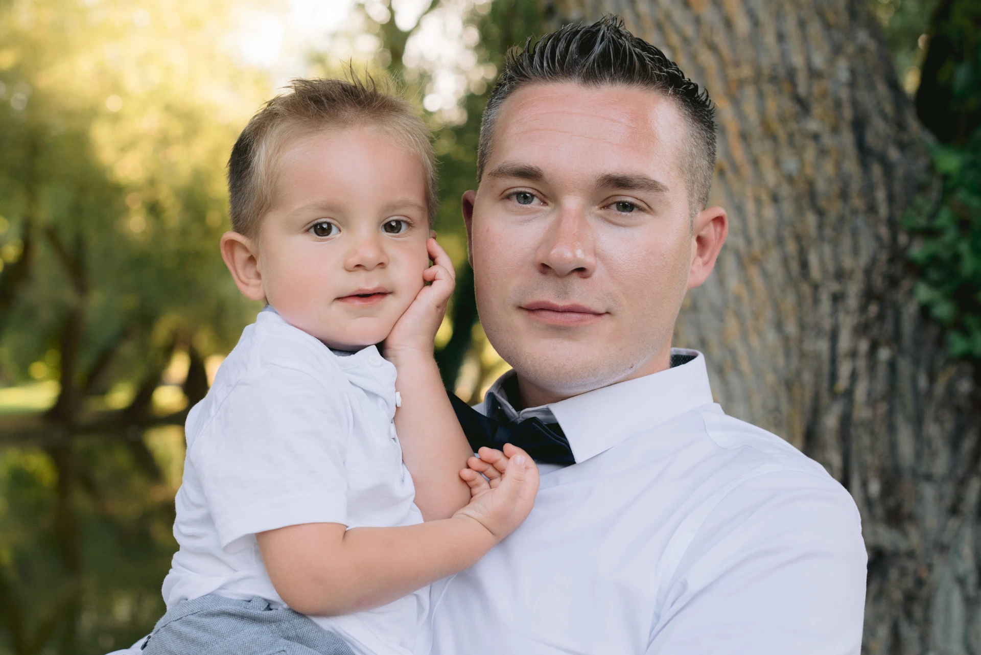 Père portant son jeune fils dans les bras, tous deux en chemise blanche, portrait de famille en extérieur par Alexandre Topolewski