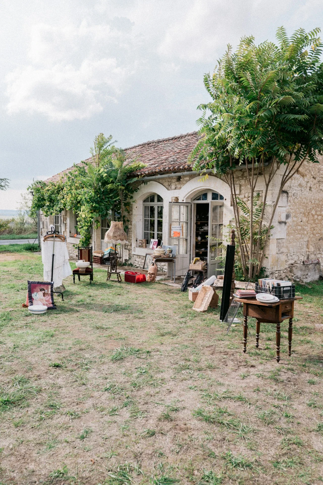 Brocante installée devant une dépendance en pierre à toit de tuiles, avec meubles, vaisselle et vêtements sur la pelouse
