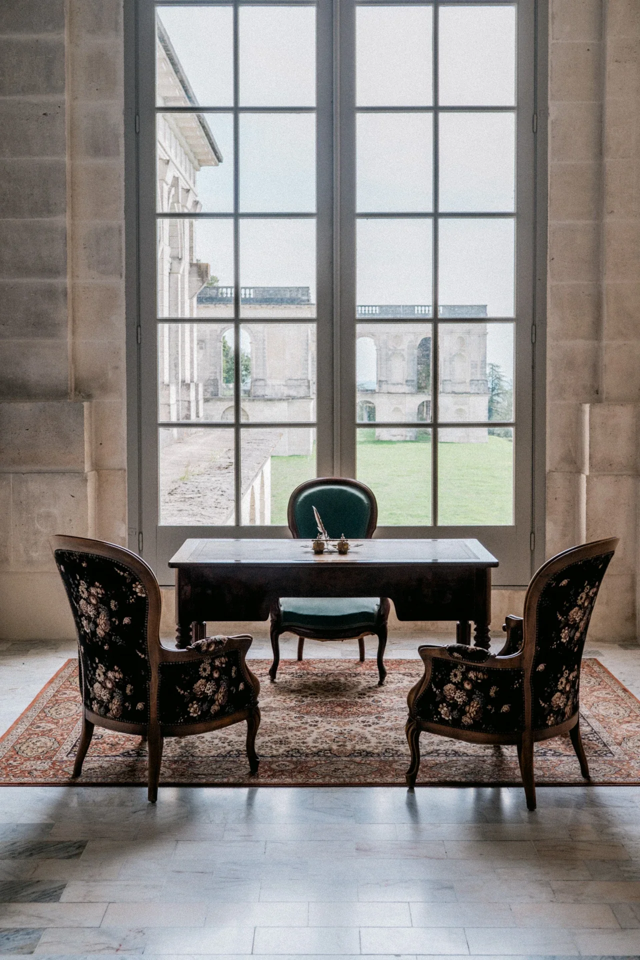 Bureau en bois et fauteuils à motifs floraux devant une haute fenêtre donnant sur les arcades du château