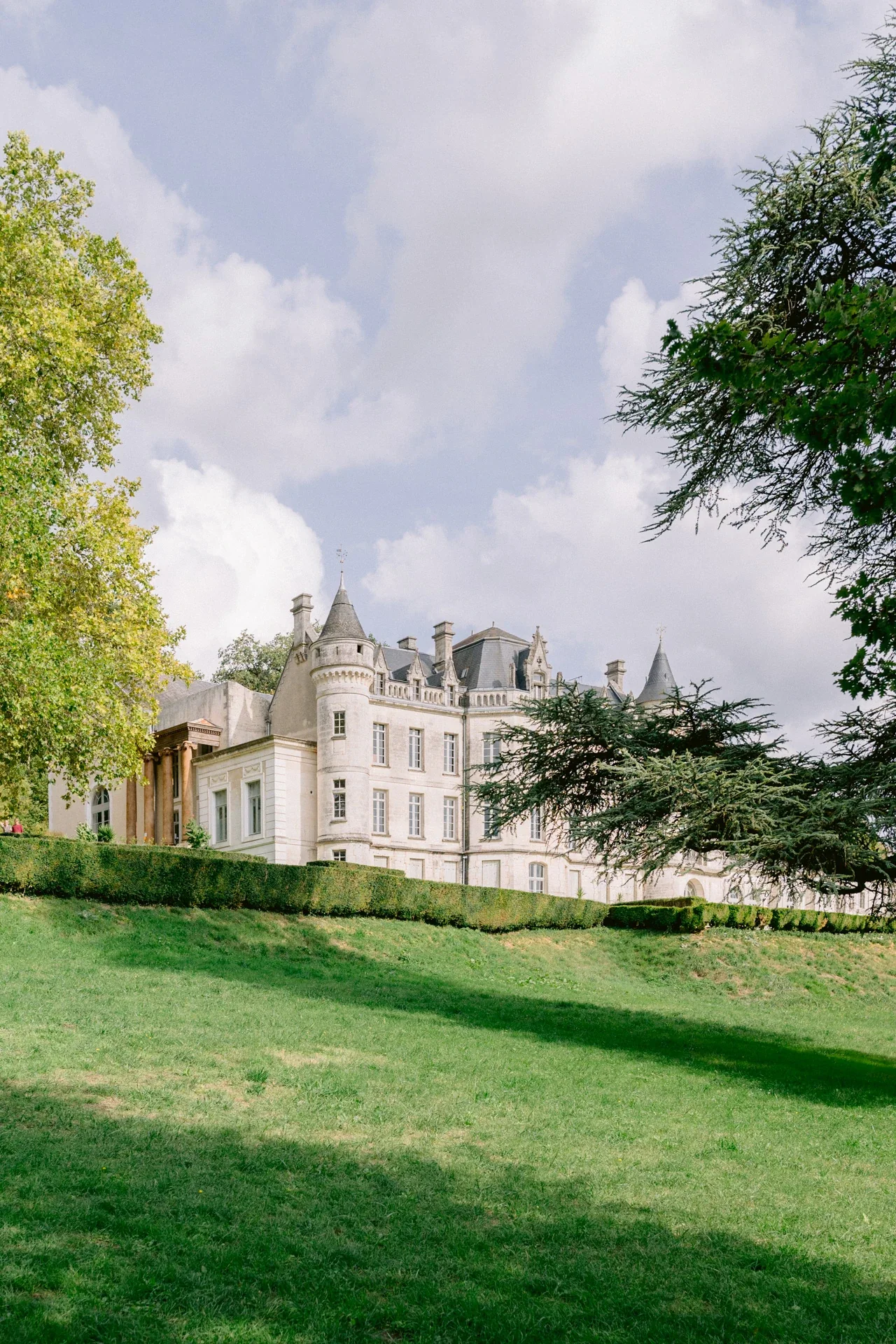 Château aux tourelles d'ardoise vu depuis la pelouse, entre un cèdre et un platane, sous un ciel de nuages