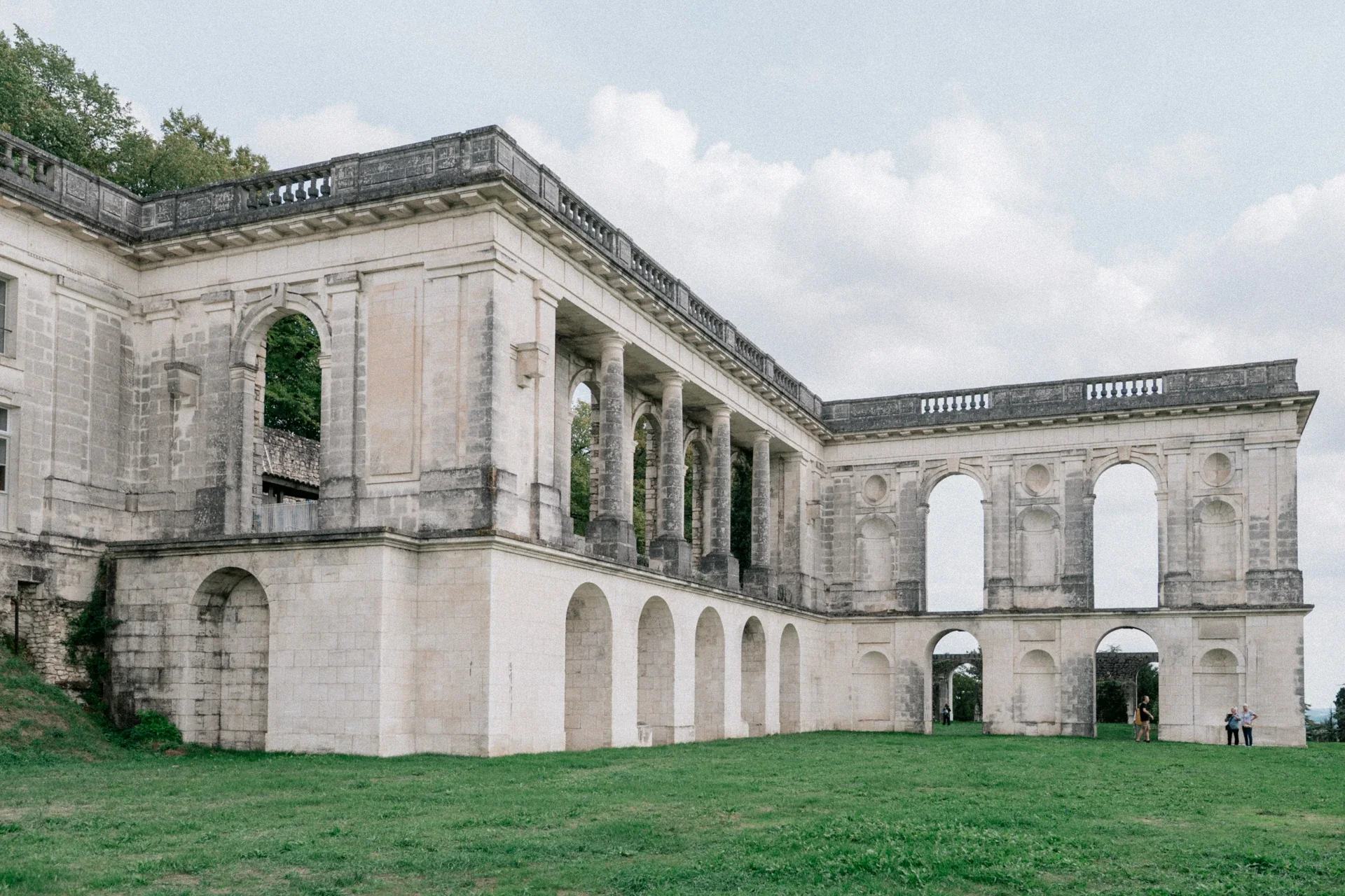 Aile à colonnade et arcades en pierre claire, avec quelques visiteurs sur la pelouse