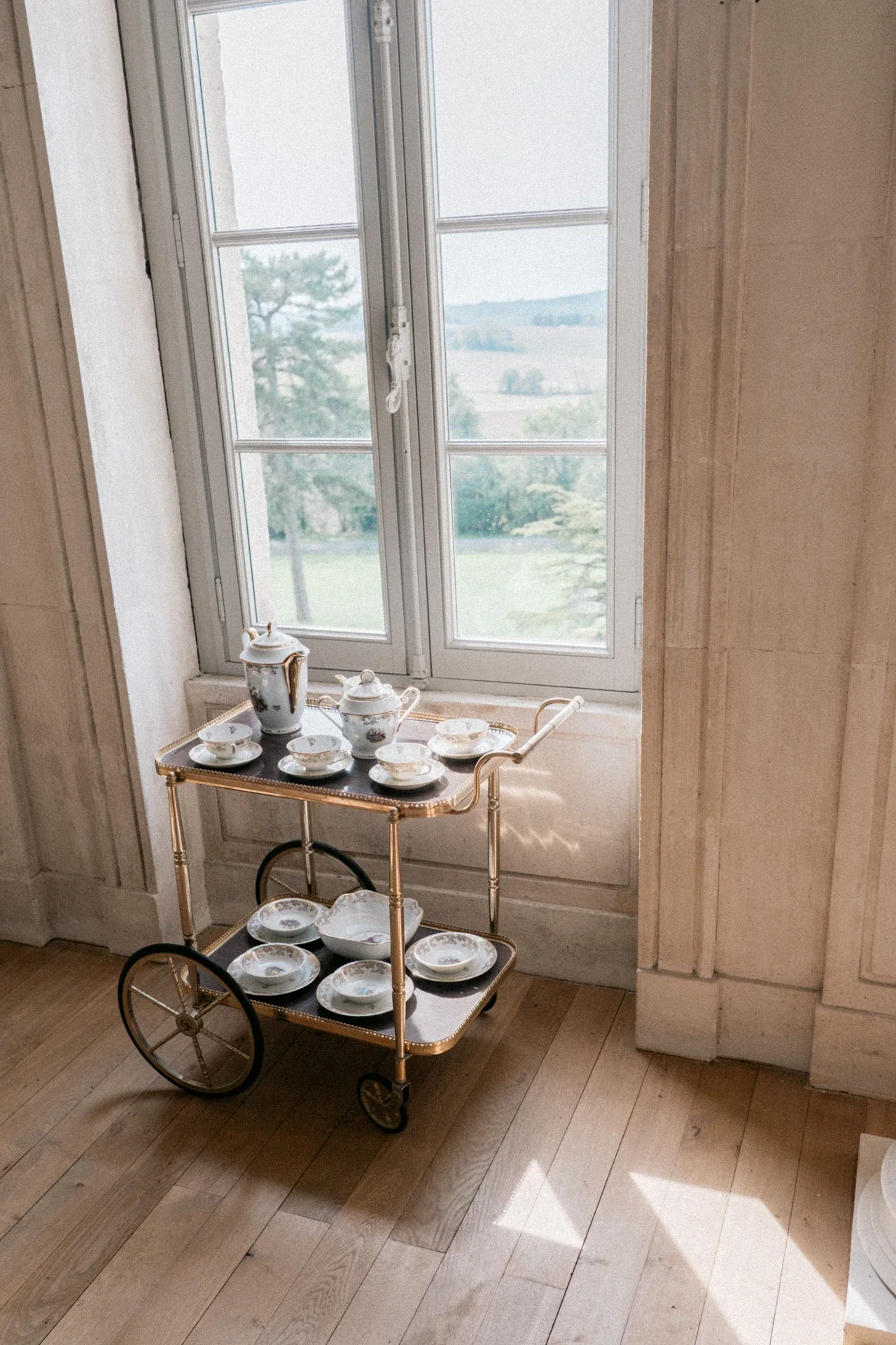 Desserte en laiton chargée d'un service à thé en porcelaine, posée près d'une fenêtre sur parquet