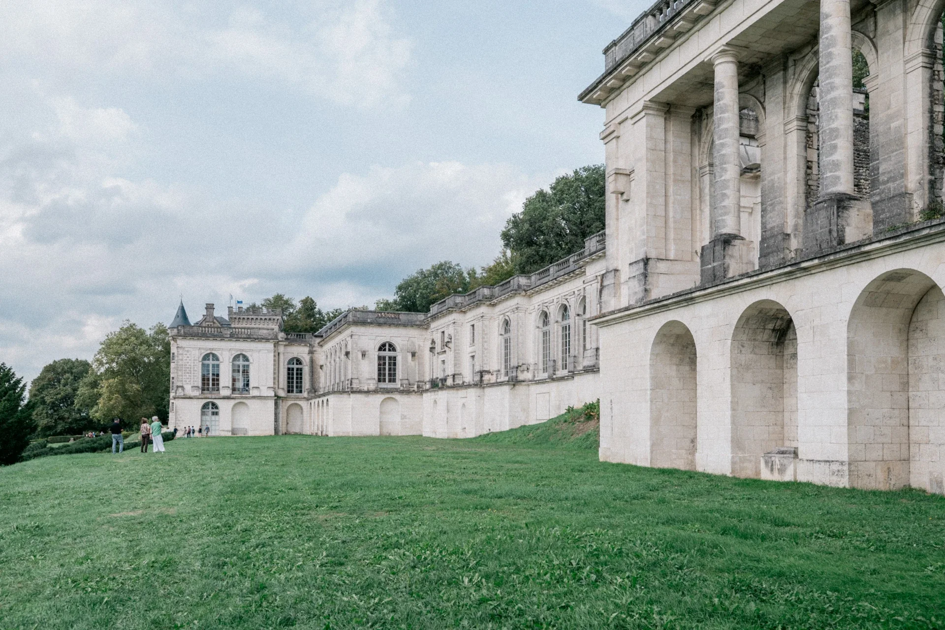 Longue façade en pierre claire du château de la Mercerie, grande pelouse et visiteurs au loin sous un ciel nuageux