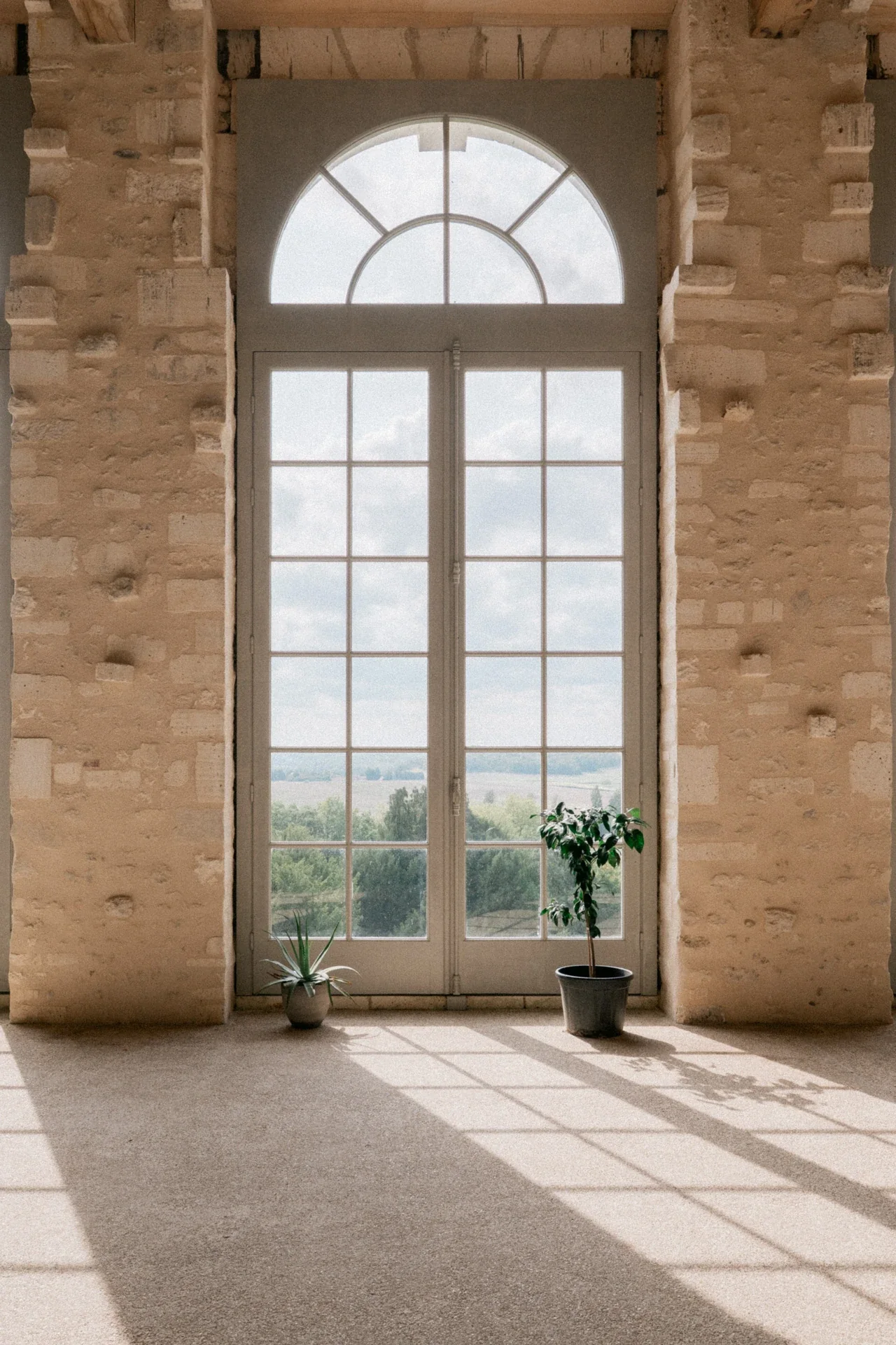 Haute fenêtre cintrée entre deux murs de pierre apparente, deux plantes en pot et ombres des carreaux sur le sol