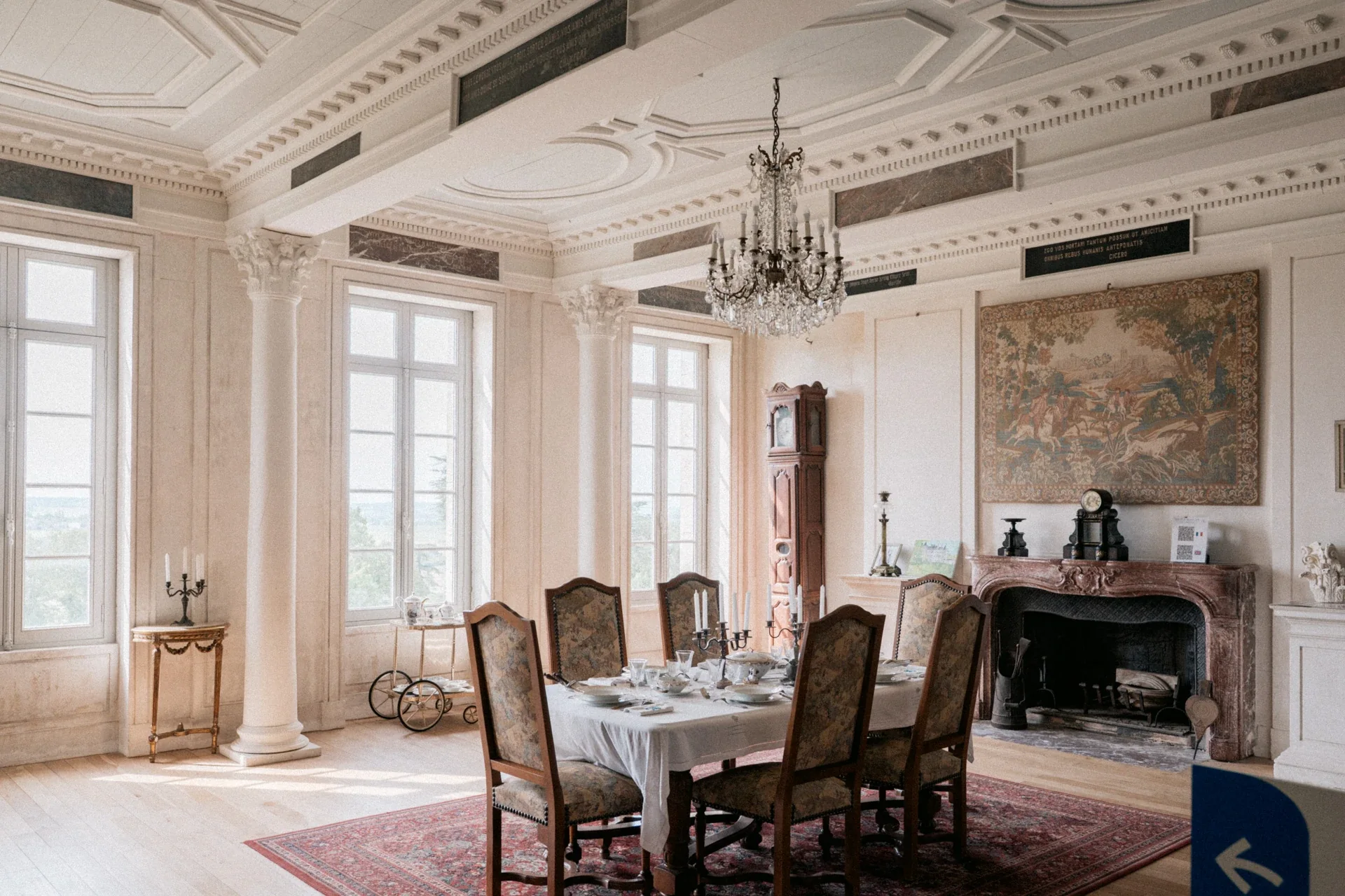 Grande salle à manger lumineuse avec table dressée, lustre en cristal, tapisserie et cheminée en marbre rose