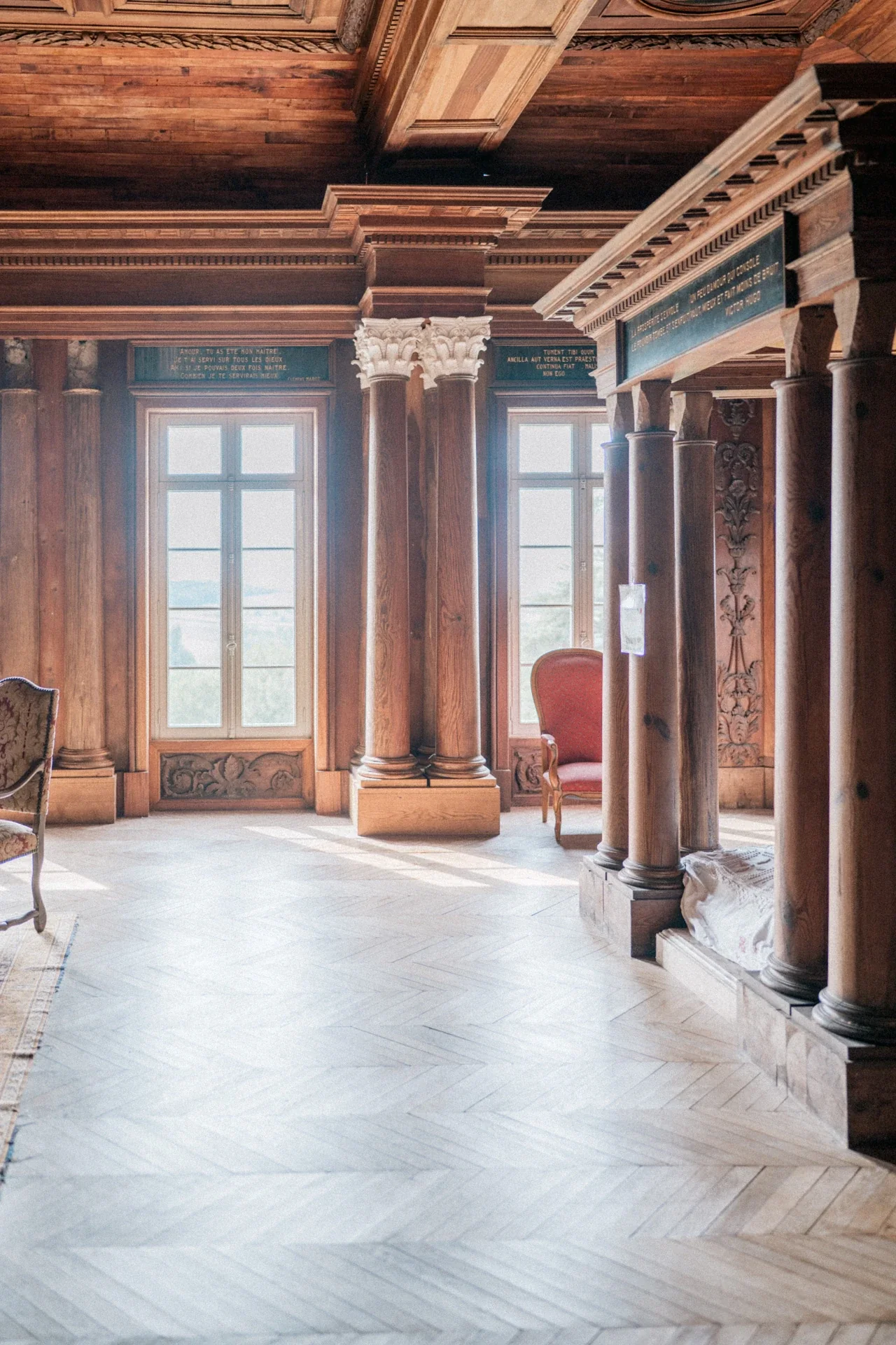 Salon en boiseries massives, colonnes de bois, grandes fenêtres et parquet à chevrons éclairé par le soleil