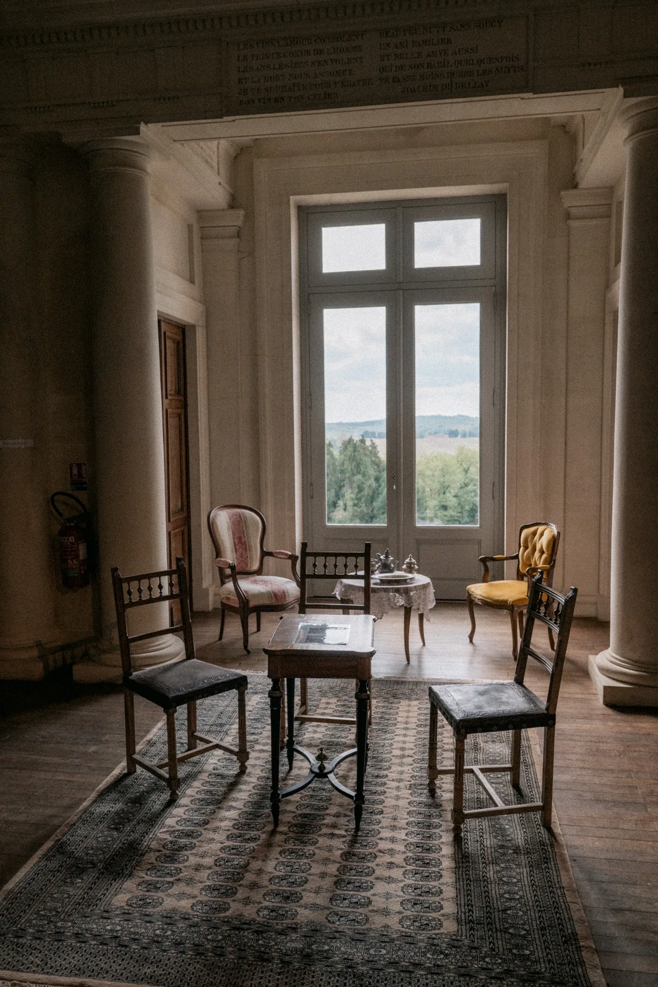 Petit salon de chaises anciennes et guéridon devant une porte-fenêtre ouverte sur la campagne