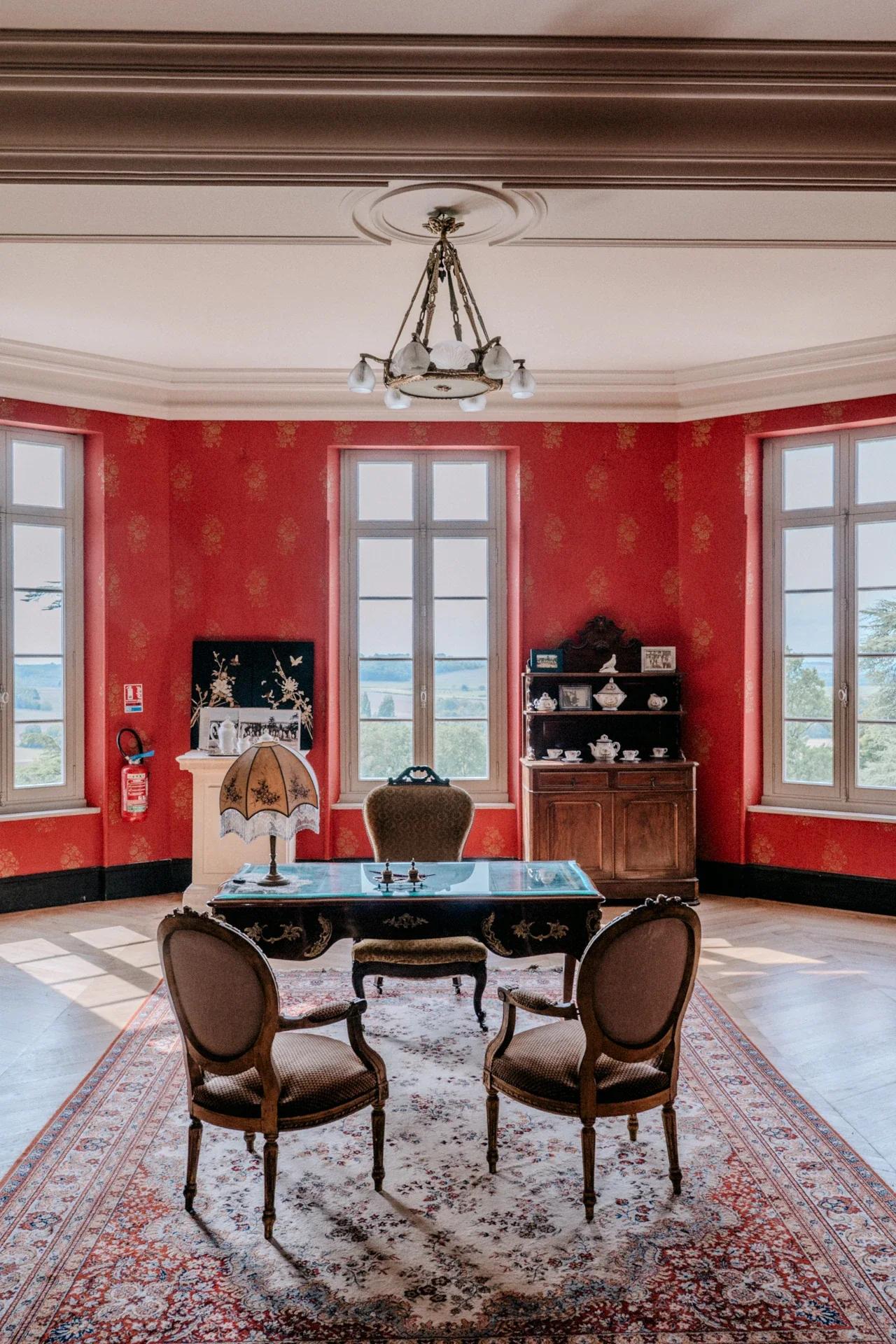 Salon aux murs rouges, bureau ancien, fauteuils médaillon et fenêtres ouvertes sur la campagne