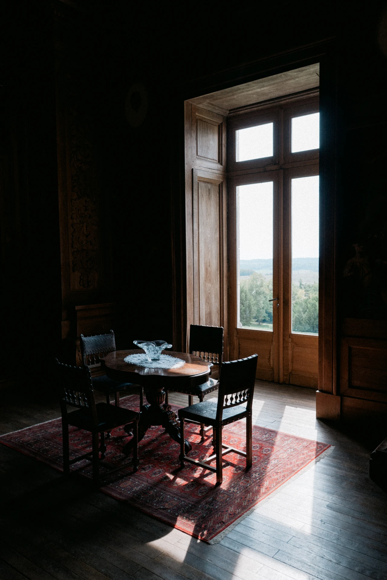 Table ronde et chaises sculptées dans une pièce sombre, éclairées par la lumière d'une porte-fenêtre