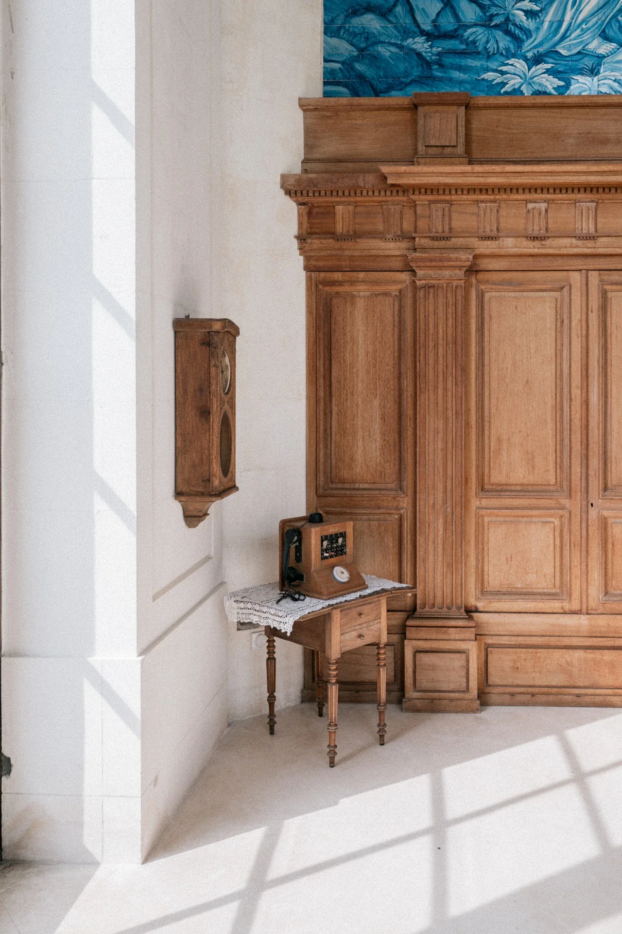 Téléphone ancien posé sur une petite table à napperon de dentelle, devant une grande boiserie en chêne et un panneau d'azulejos bleus