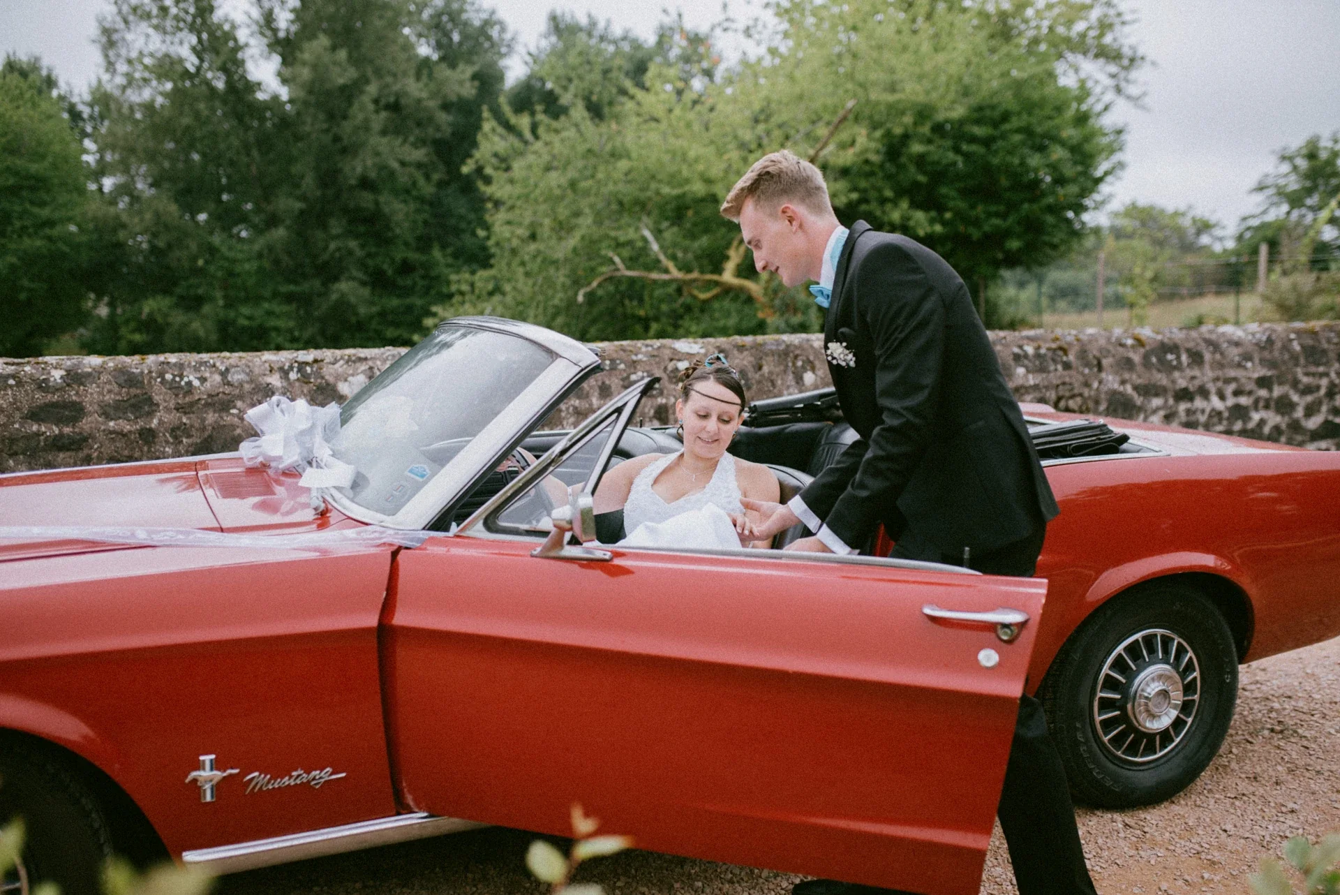 Le marié penché au-dessus de la portière ouverte de la Mustang pour aider la mariée à s’installer