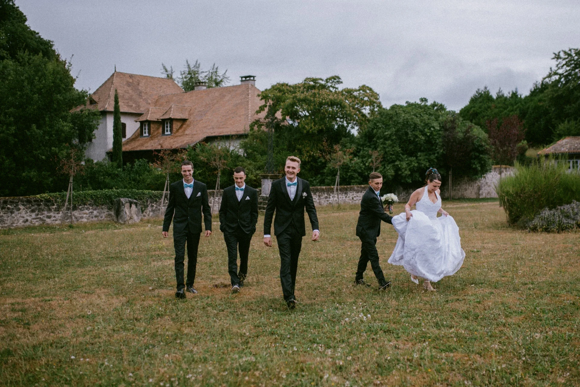 Le marié et deux témoins avançant en souriant sur la pelouse, un autre témoin aidant la mariée à tenir sa robe