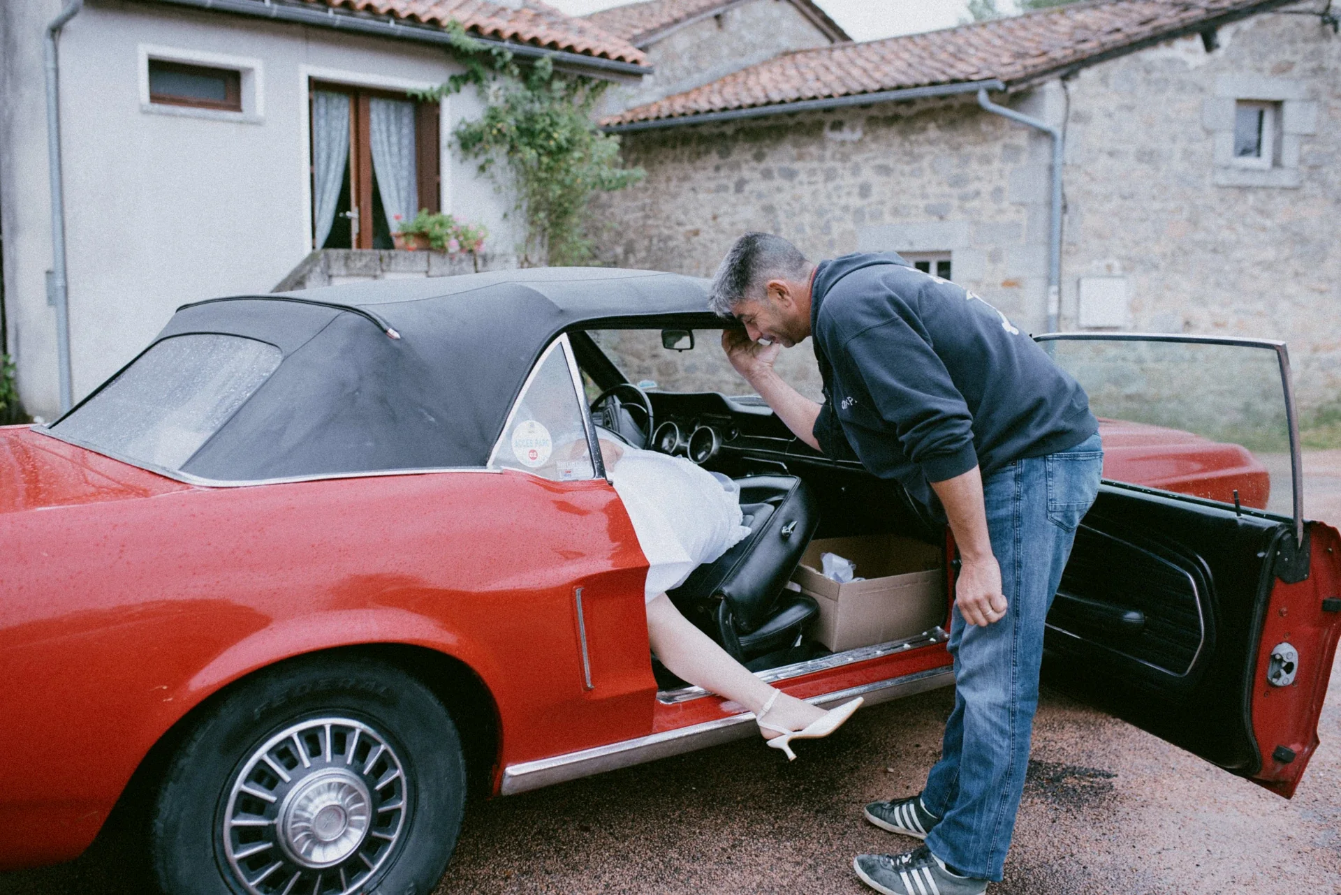 Mariée en robe blanche et escarpins montant dans la Mustang rouge sous la pluie, aidée par un proche penché vers l’habitacle
