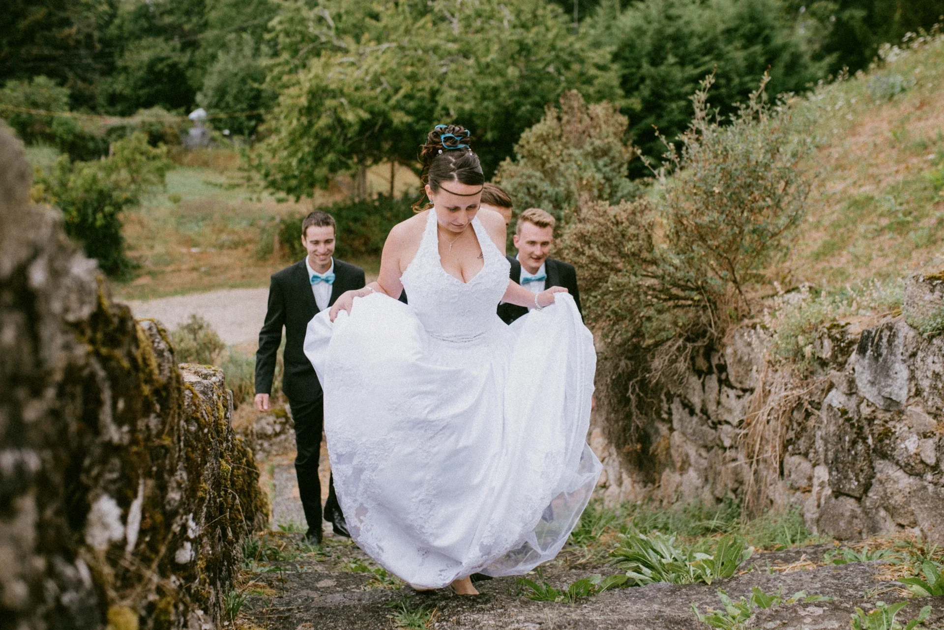 Mariée relevant sa robe pour monter un escalier de pierre moussue, suivie de deux témoins