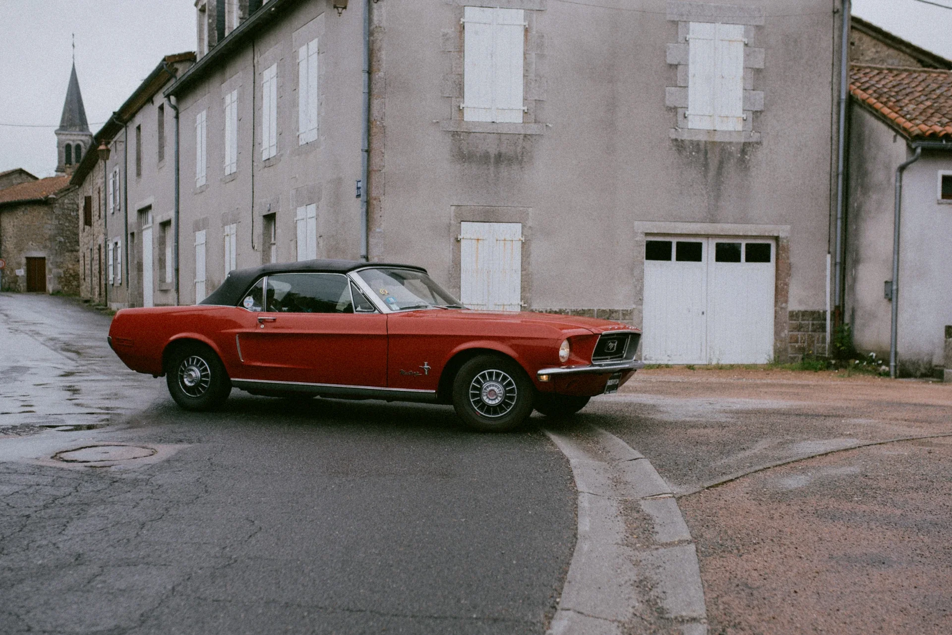 Ford Mustang rouge décapotable tournant au coin d’une rue de Piégut-Pluviers, jour de pluie
