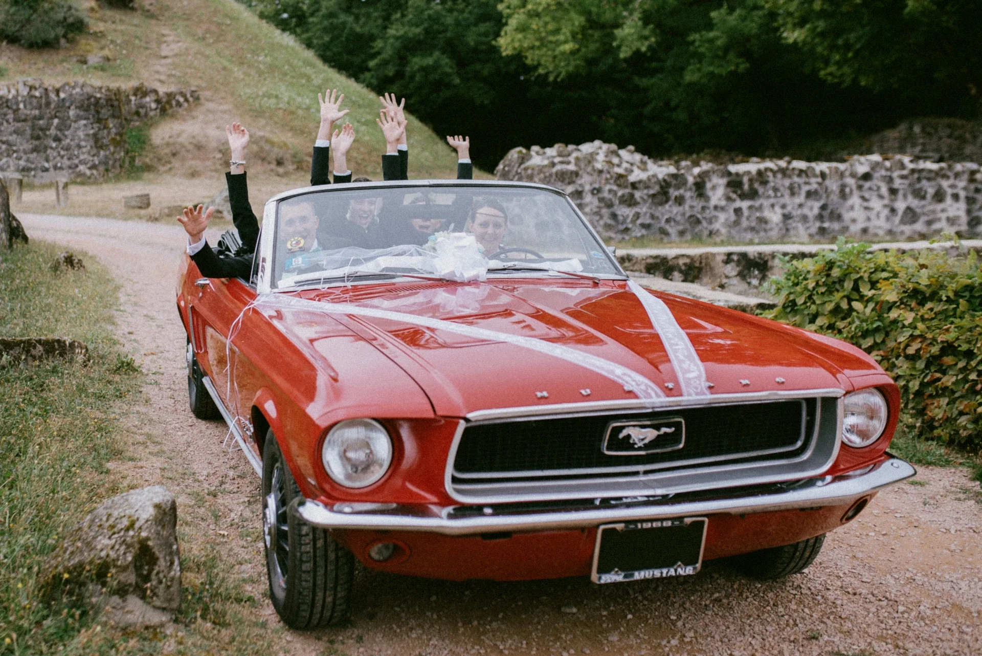 La Mustang rouge ornée de rubans blancs avançant sur le chemin des ruines, les témoins levant les bras à l’arrière