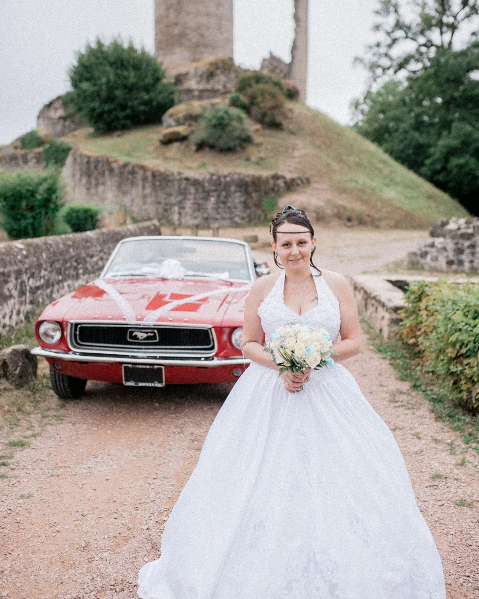 Portrait de la mariée en robe blanche tenant son bouquet, devant la Mustang rouge et la butte du château