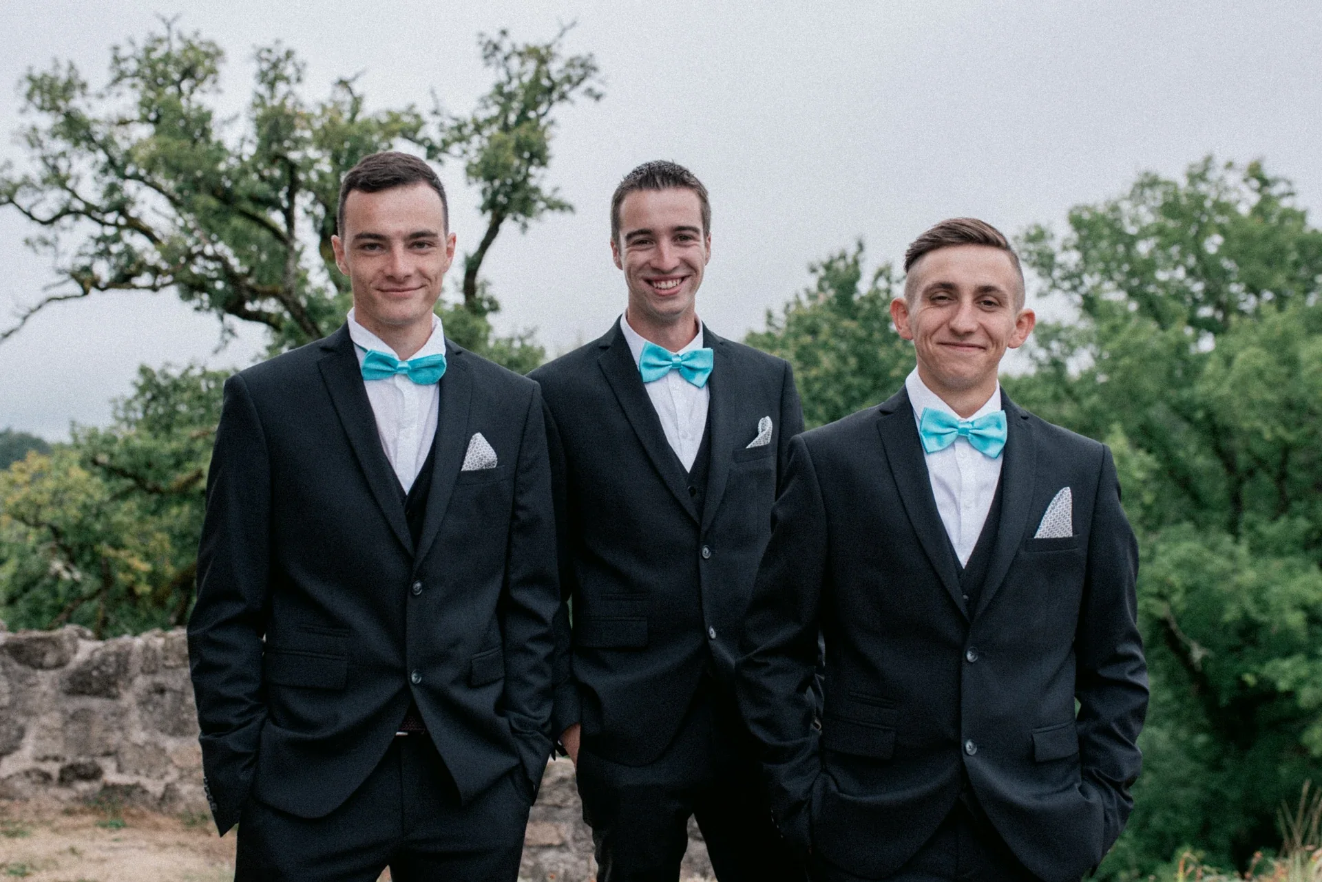 Portrait des trois témoins souriants, mains dans les poches, en costume sombre et nœud papillon turquoise