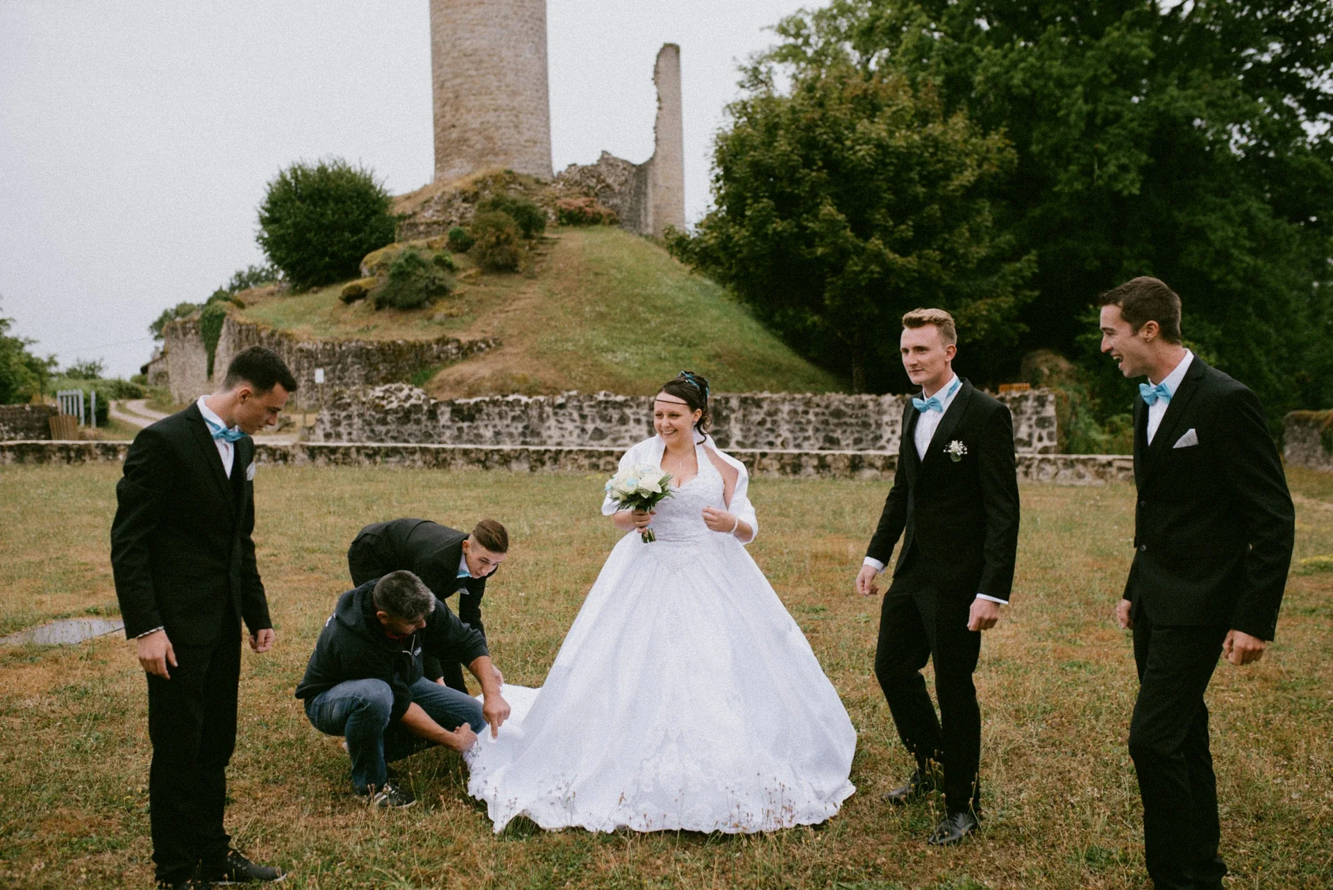 Mariée rieuse devant la tour en ruines pendant que deux témoins ajustent le bas de sa robe dans l’herbe, entourée de deux autres témoins