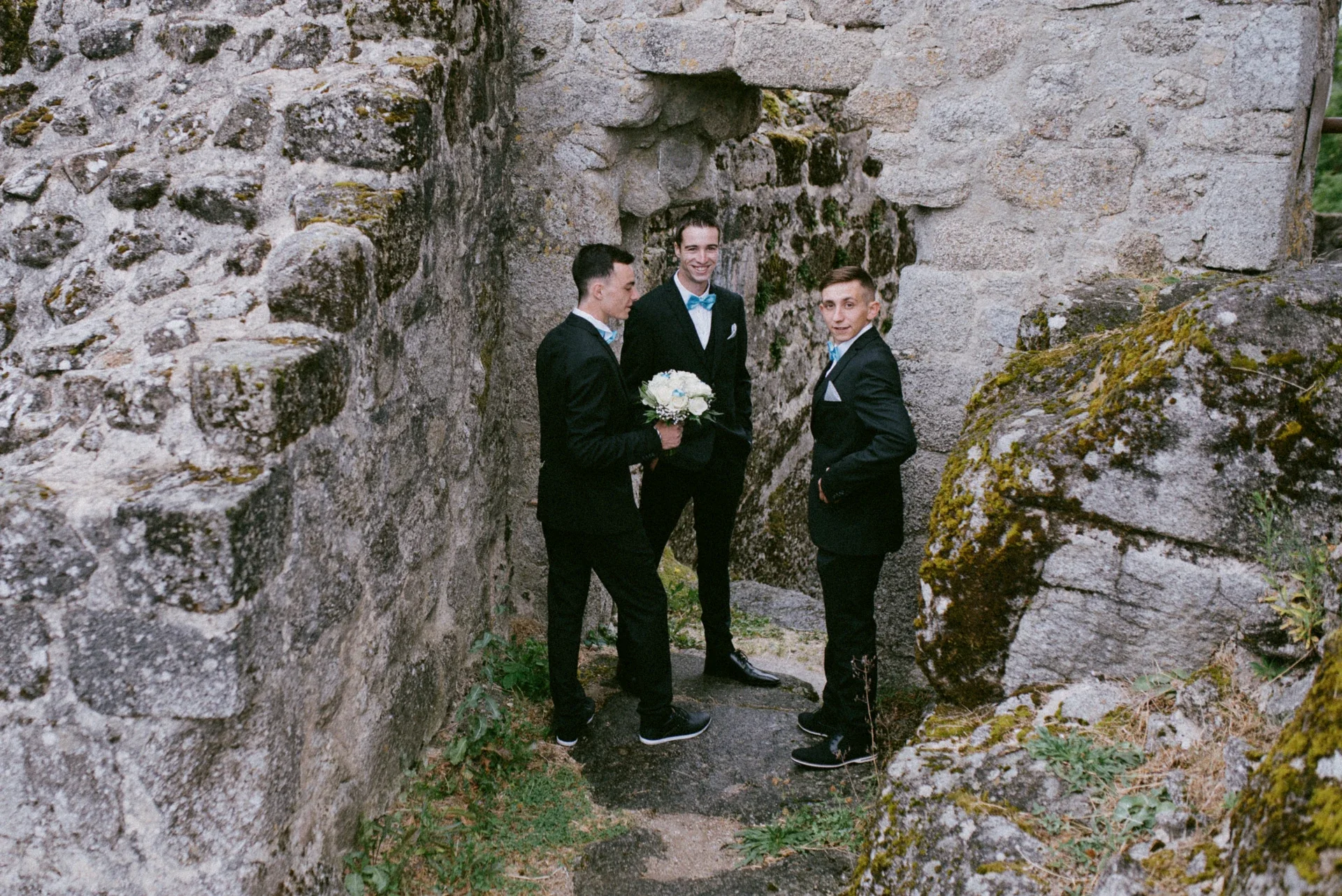 Trois témoins en costume noir et nœud papillon turquoise discutant dans un passage de pierre, l’un tenant le bouquet de la mariée