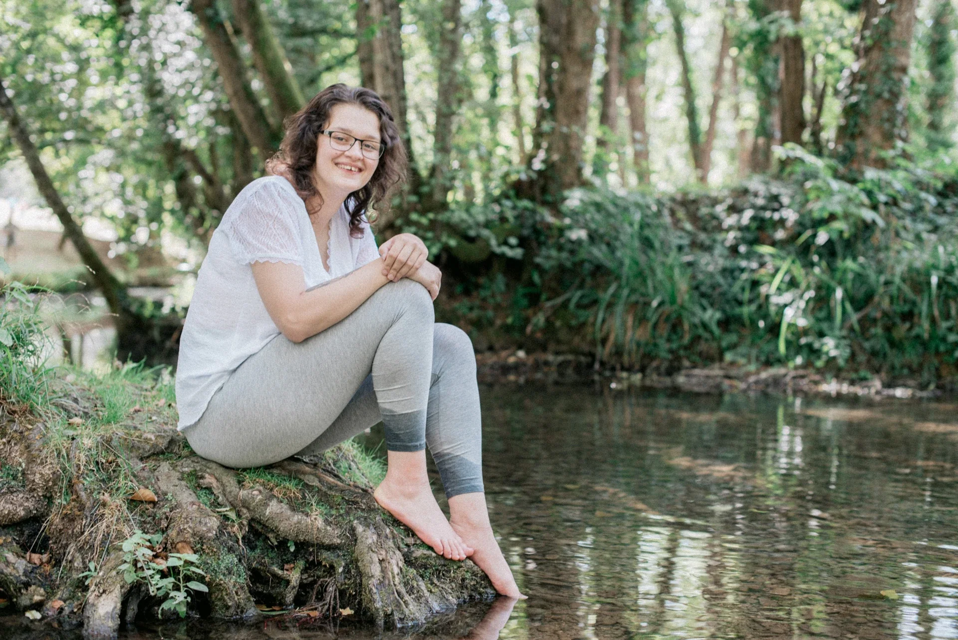 Jeune femme assise sur les racines d'un arbre au bord de l'eau, les orteils effleurant la surface, sous les arbres du Lac du Causse