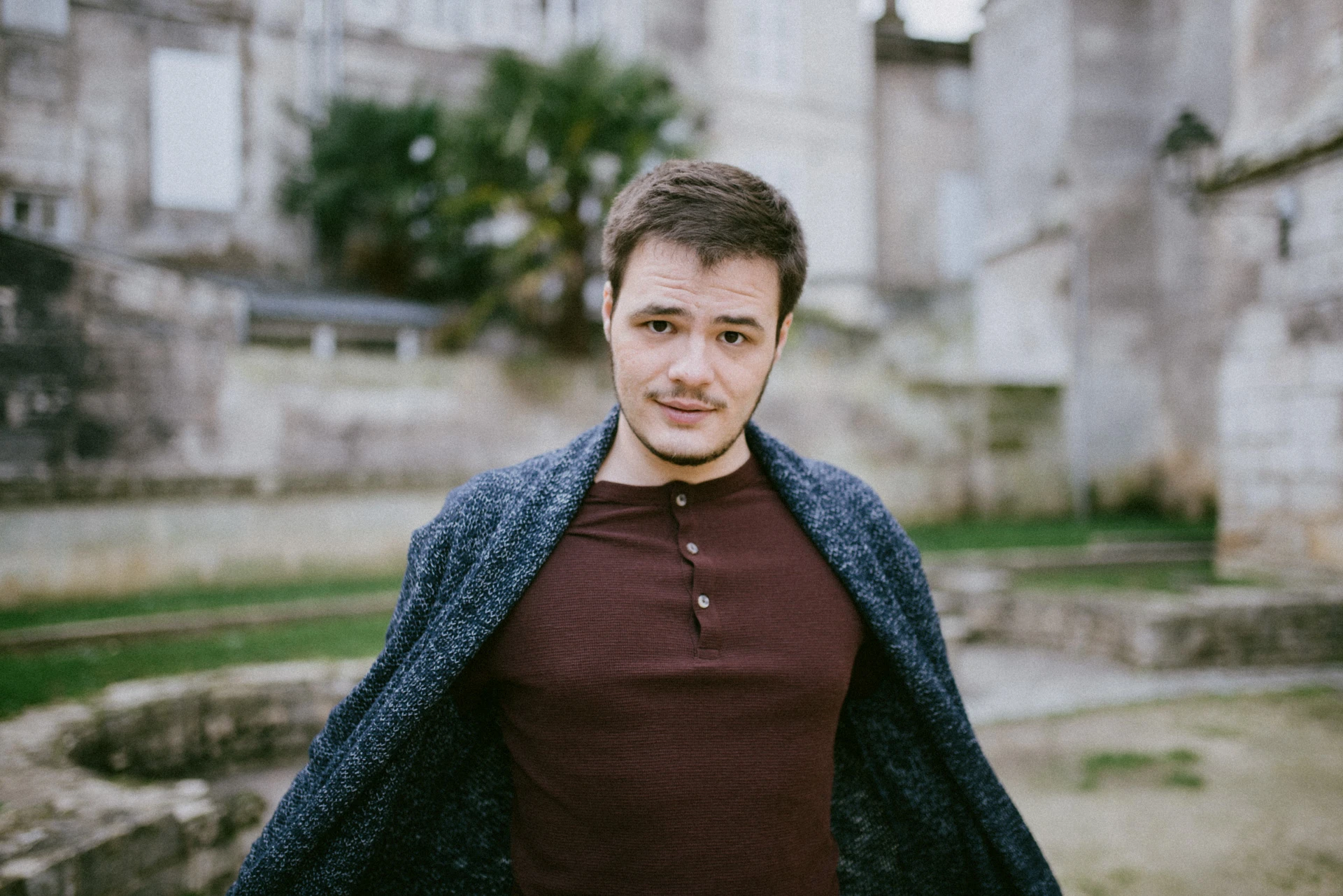 Portrait frontal d'un comédien, gilet bleu chiné ouvert sur un t-shirt bordeaux, devant des murs de pierre et un jardin