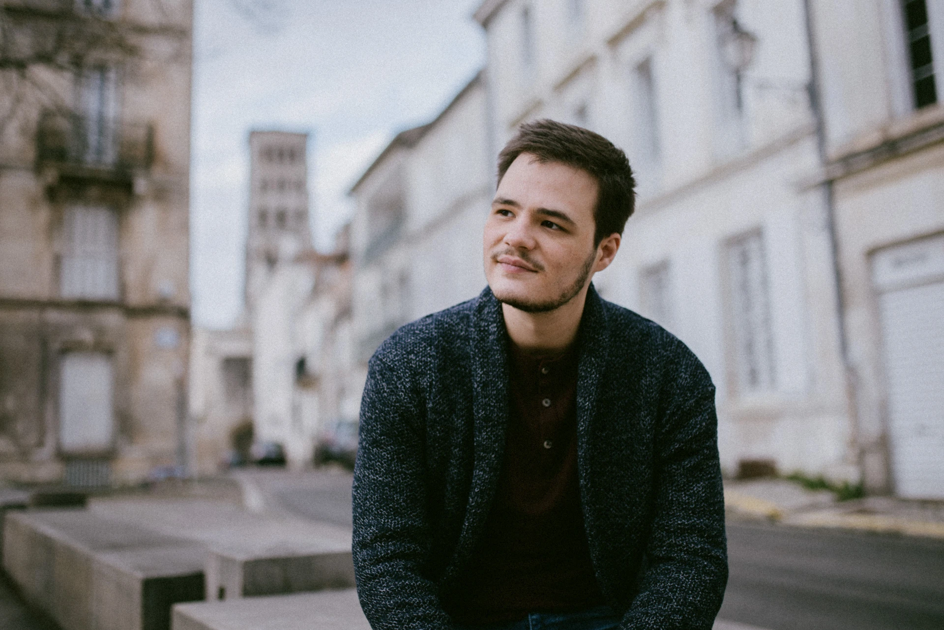 Portrait d'un comédien en gilet bleu chiné et t-shirt bordeaux, assis dans une rue d'Angoulême, un sourire au coin des lèvres