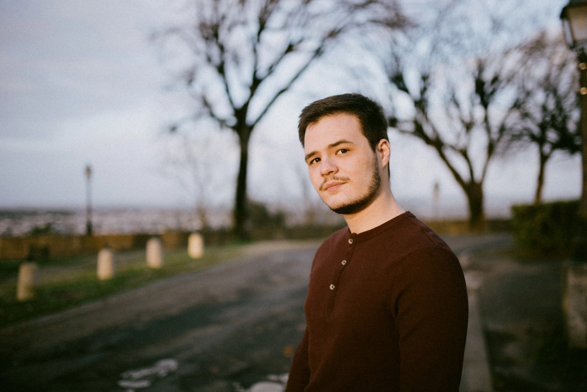 Portrait d'un comédien en pull bordeaux à la lumière dorée du soir, devant des arbres nus et la vue sur la ville d'Angoulême