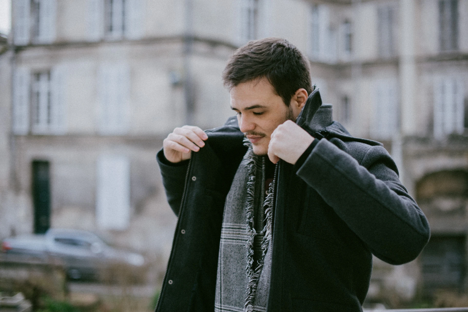Portrait d'un comédien ajustant le col de son manteau sombre, écharpe à carreaux autour du cou, devant une façade en pierre