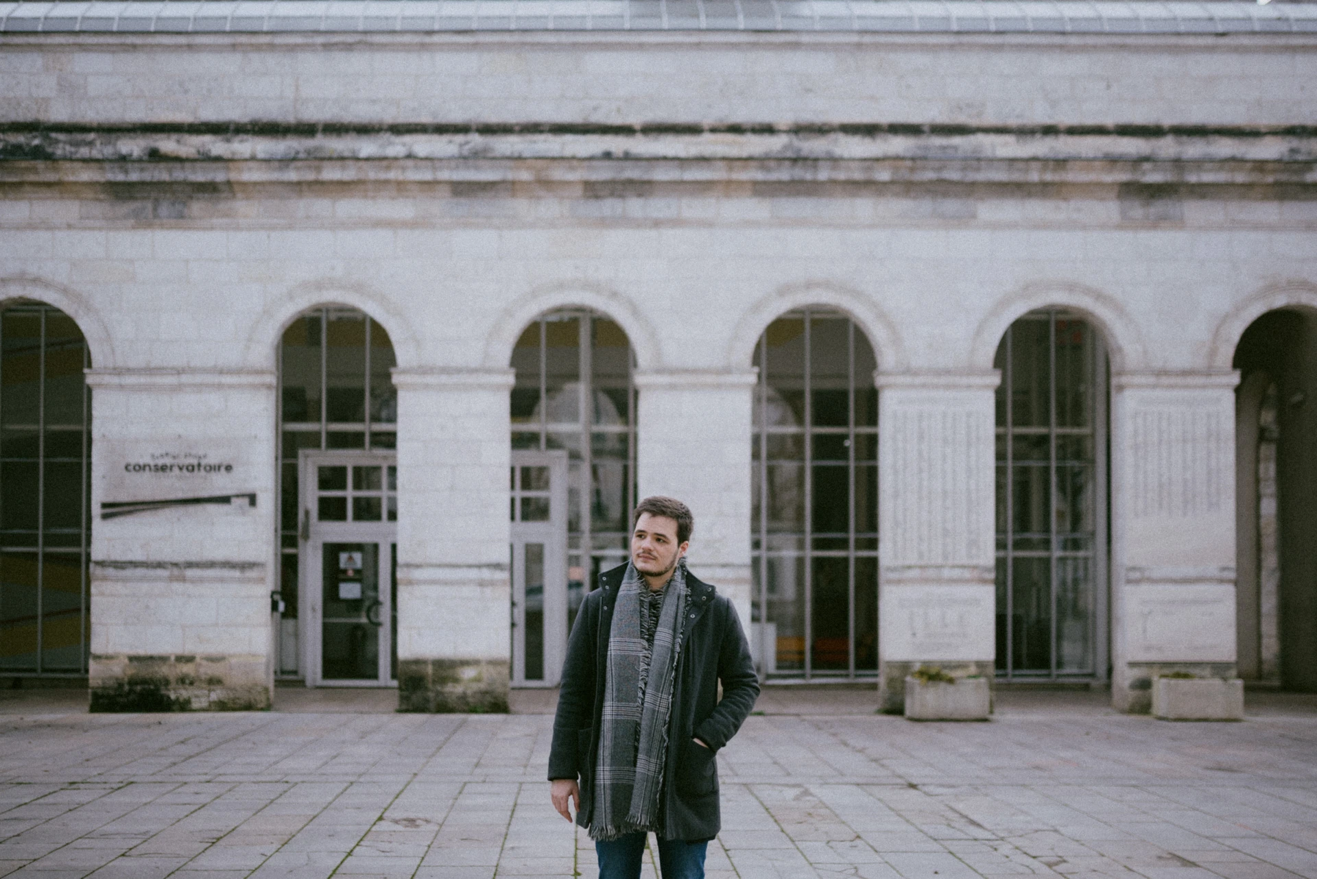 Portrait d'un comédien en manteau et écharpe, debout sur un parvis devant les arcades du conservatoire d'Angoulême