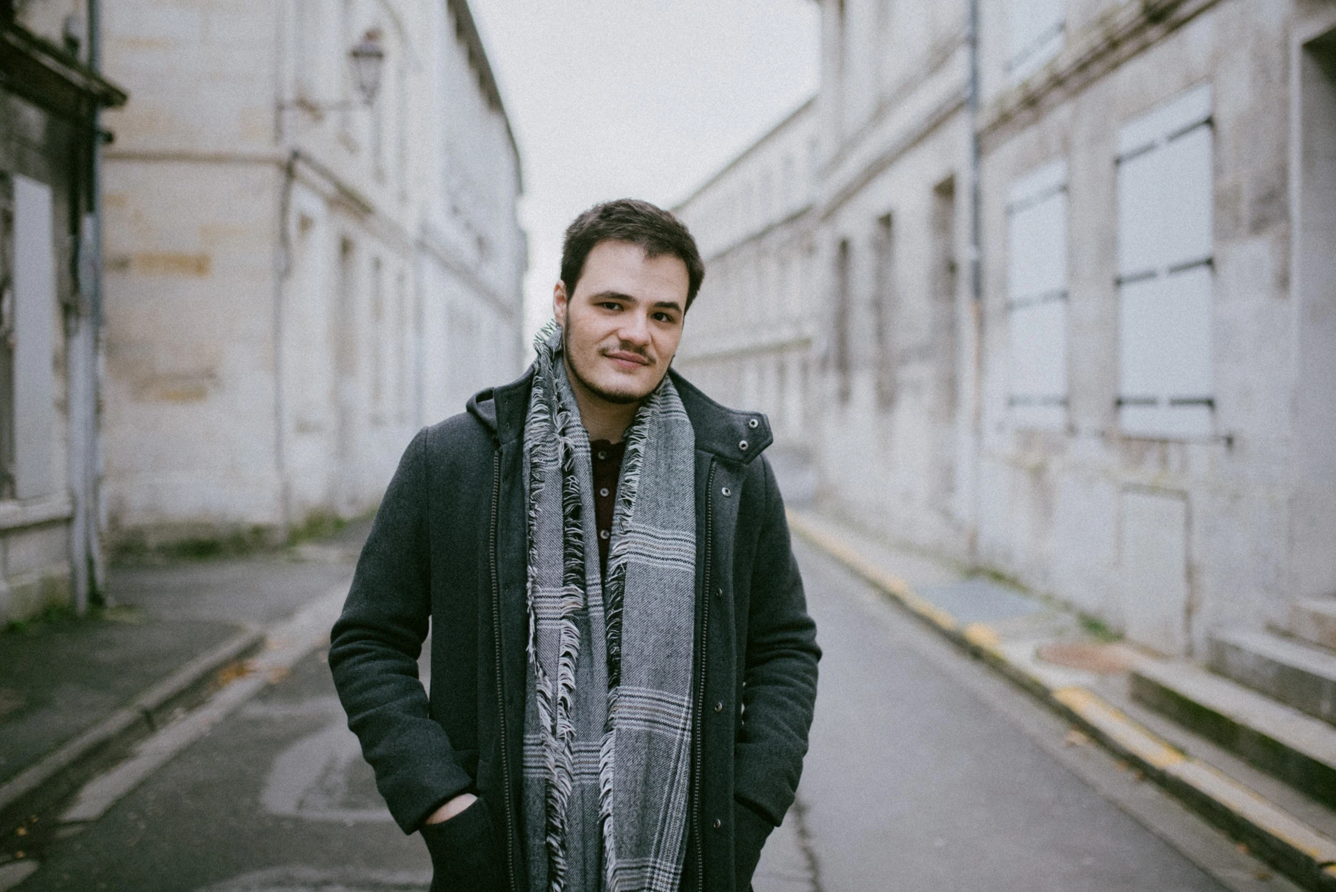 Portrait d'un comédien, mains dans les poches de son manteau, écharpe à carreaux, au milieu d'une rue bordée de façades en pierre