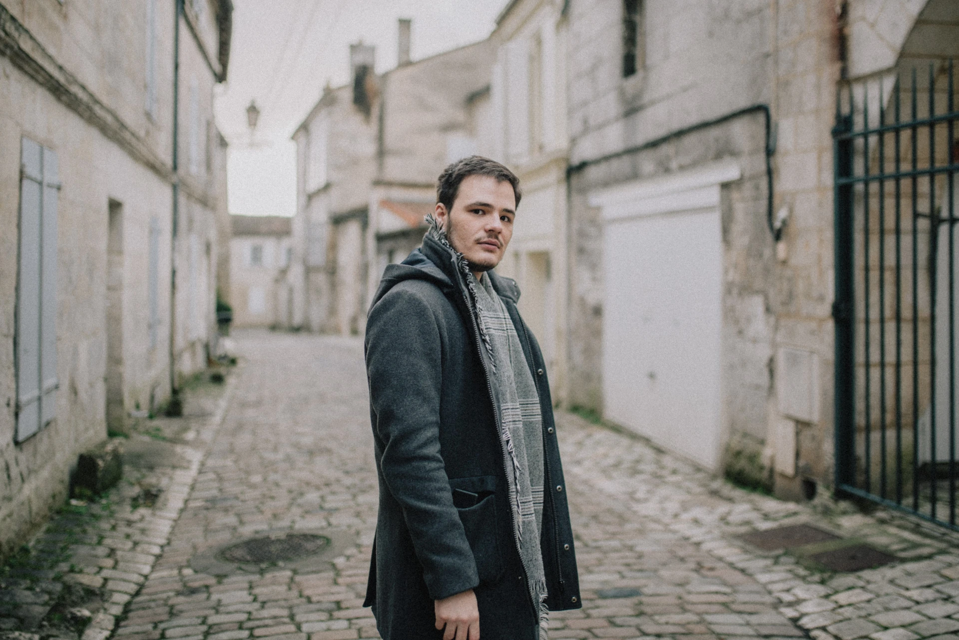 Portrait d'un comédien se retournant dans une ruelle pavée d'Angoulême, manteau gris et écharpe à carreaux