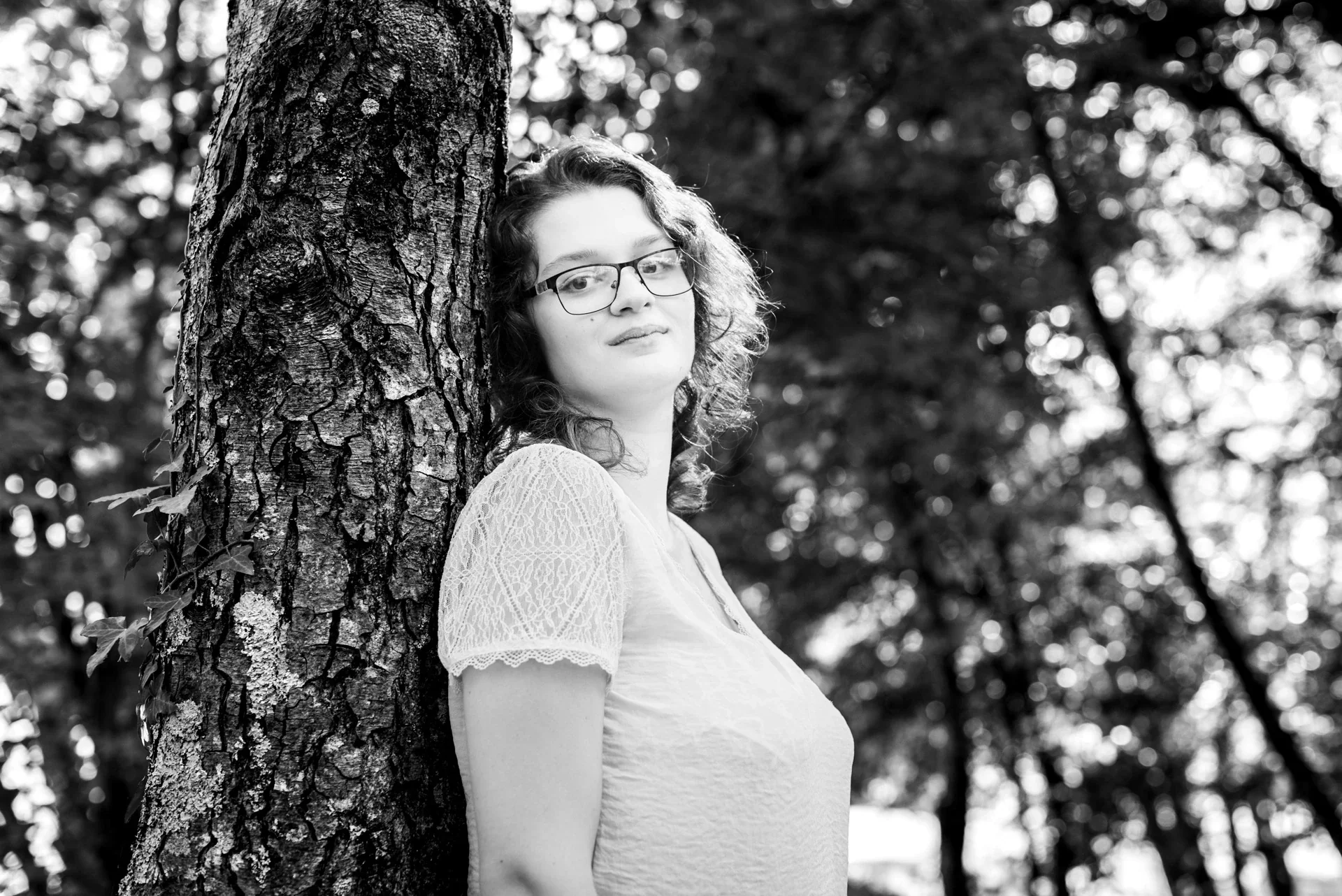 Portrait en noir et blanc d'une jeune femme adossée à un tronc d'arbre, fond de feuillages en contre-jour au Lac du Causse