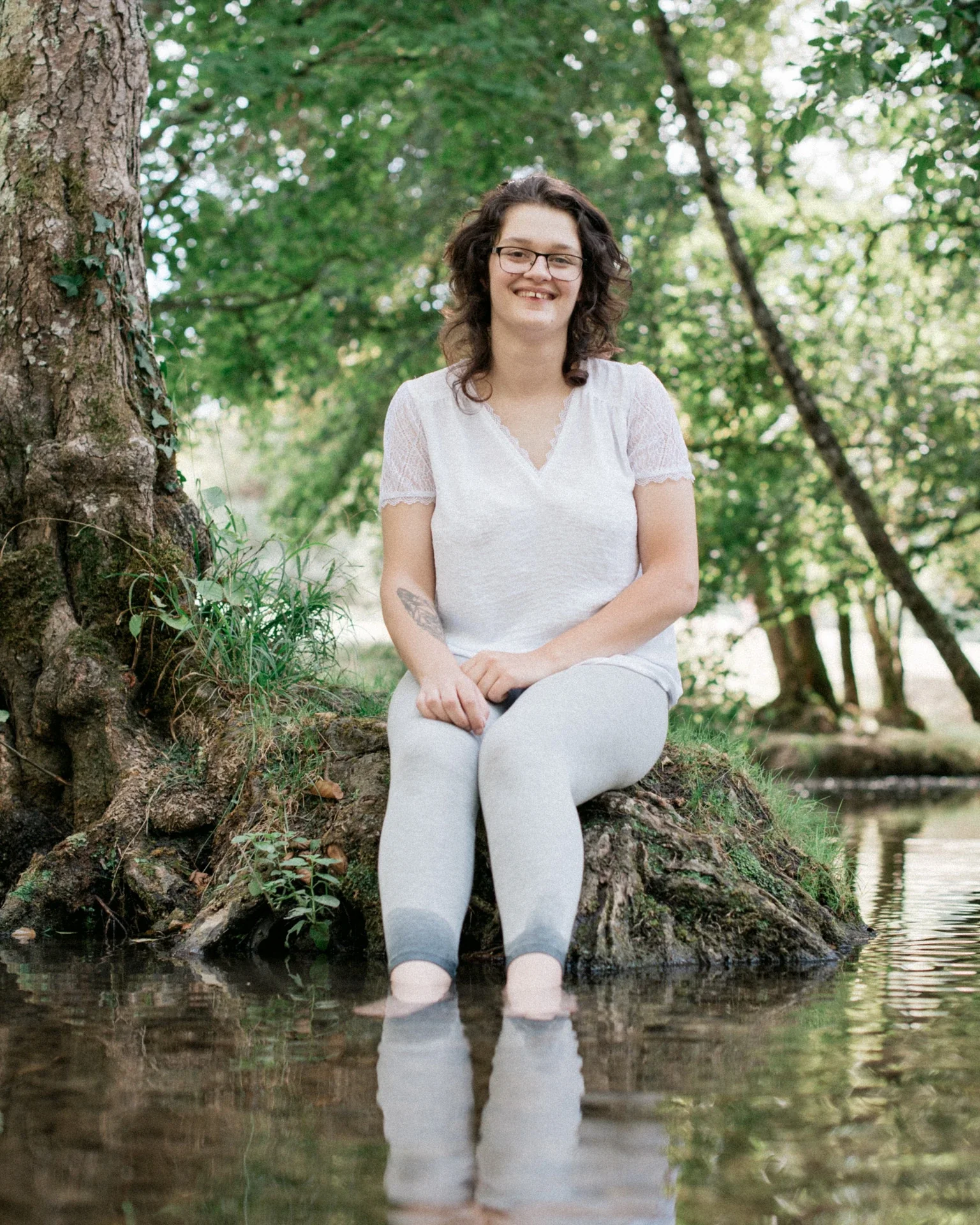 Portrait d'une jeune femme souriante assise sur une souche moussue, les pieds dans l'eau, au pied d'un grand arbre au Lac du Causse en Corrèze