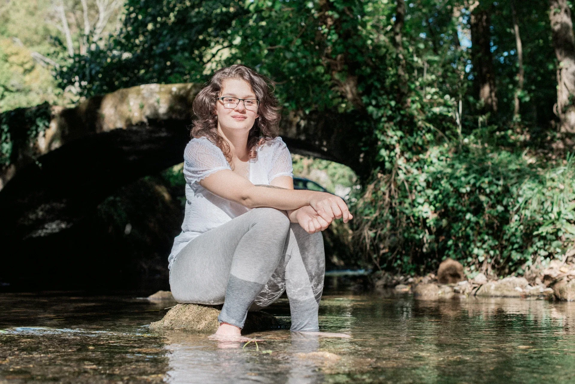 Portrait d'une jeune femme assise sur un rocher dans l'eau, devant un vieux pont de pierre couvert de lierre au Lac du Causse