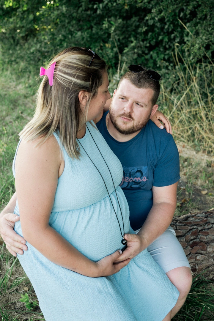 Futur papa assis contre sa compagne enceinte, la main posée sur son ventre, sur un tronc en bord de vigne