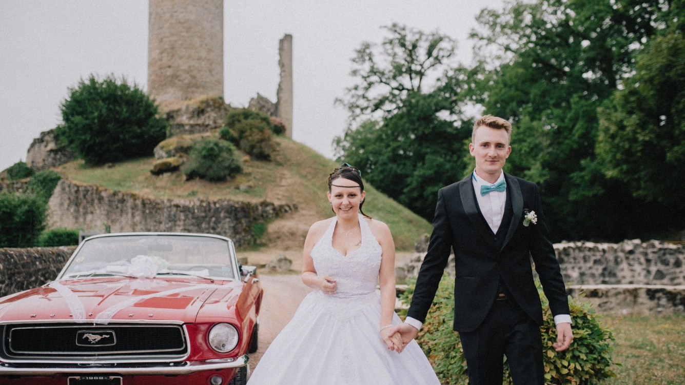 reportage de photographie de mariage en Dordogne. Un couple de mariés heureux se tient la main devant une Ford Mustang rouge, avec une tour médiévale en arrière-plan. La mariée porte une robe blanche élégante, tandis que le marié est en costume noir avec un nœud papillon turquoise. La scène se déroule dans un cadre naturel verdoyant, apportant un charme romantique à la photo