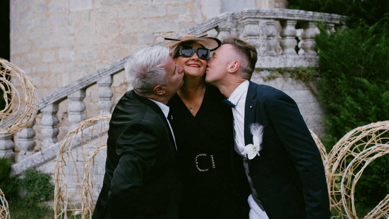 Moment de complicité capturé lors d'un reportage photo de mariage en Charente : deux hommes en costume embrassent une femme élégamment vêtue de noir, souriante et portant des lunettes de soleil, dans un cadre extérieur raffiné avec des escaliers en pierre en arrière-plan. Cette scène chaleureuse reflète l’ambiance conviviale et festive de l'événement, immortalisée dans un style Fine Art.