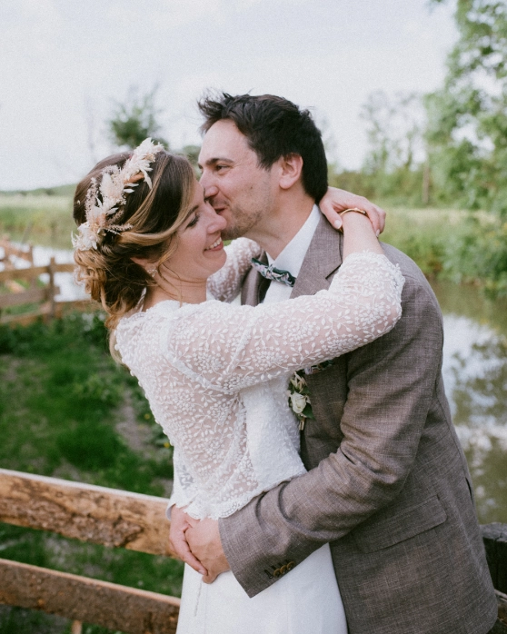 Un couple de mariés heureux partage un moment de tendresse en extérieur. La mariée porte une robe en dentelle et une couronne florale, tandis que le marié est habillé d’un costume beige et d’un nœud papillon. Ils se tiennent sur un pont en bois près d’un paysage bucolique.