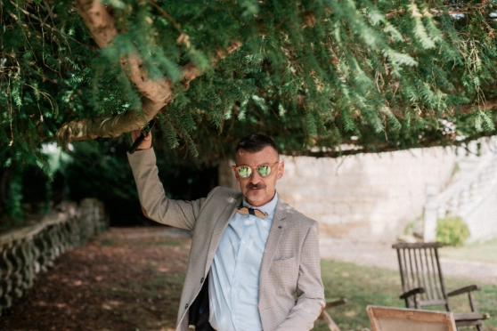 Un homme élégant portant des lunettes de soleil réfléchissantes, un costume beige à carreaux et un nœud papillon en bois. Il est debout sous un arbre dans un cadre extérieur verdoyant, posant avec assurance tout en tenant une branche.