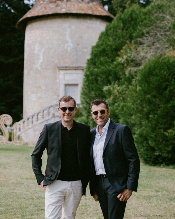 Deux hommes élégants, vêtus de costumes sombres et portant des lunettes de soleil, posent dans un cadre extérieur verdoyant. En arrière-plan, on distingue une tourelle en pierre et des buissons bien entretenus.