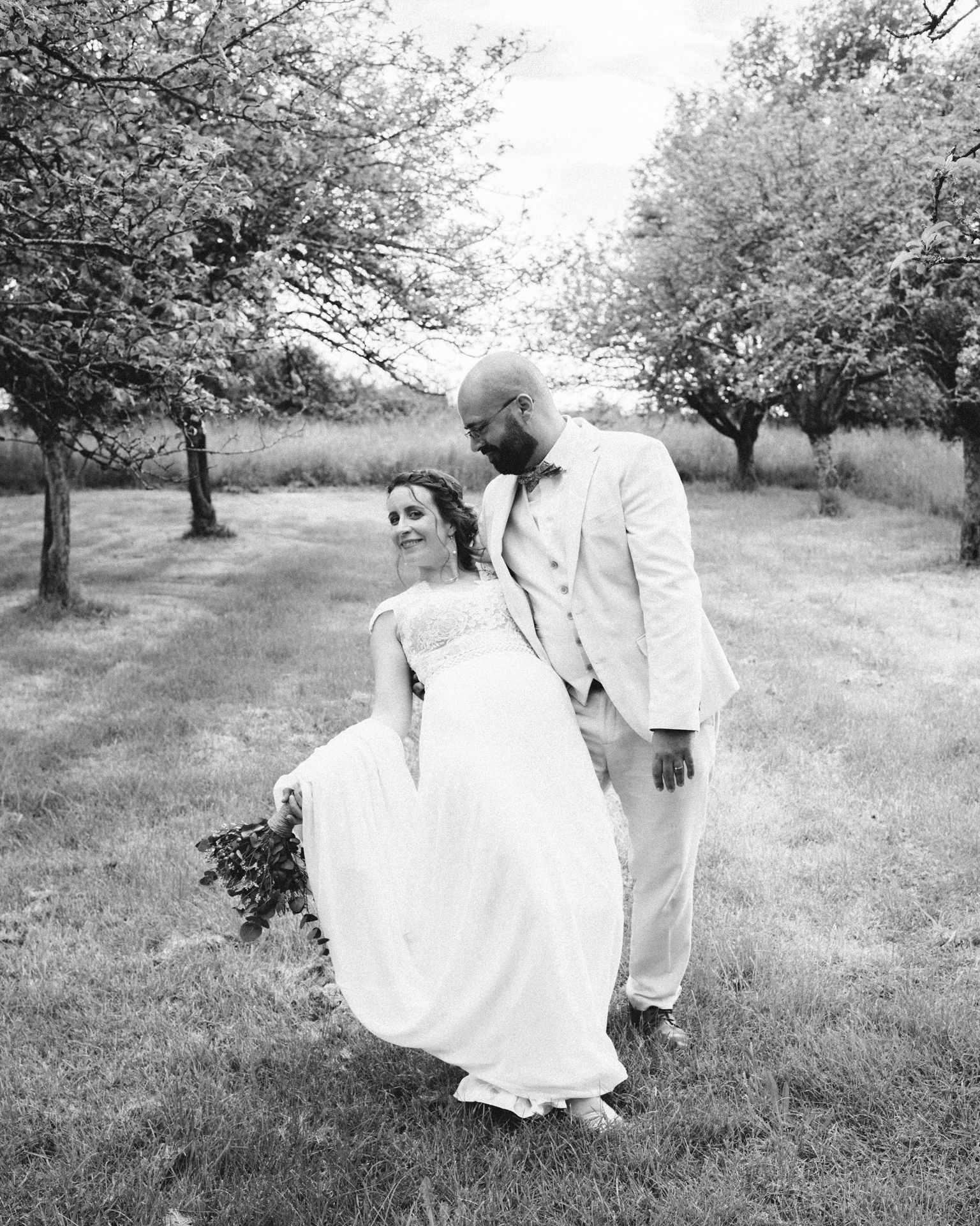 Mariés élégants dans un verger, le marié en costume clair regarde tendrement la mariée en robe fluide, bouquet à la main – photographie artistique en noir et blanc prise en Charente ou Gironde.