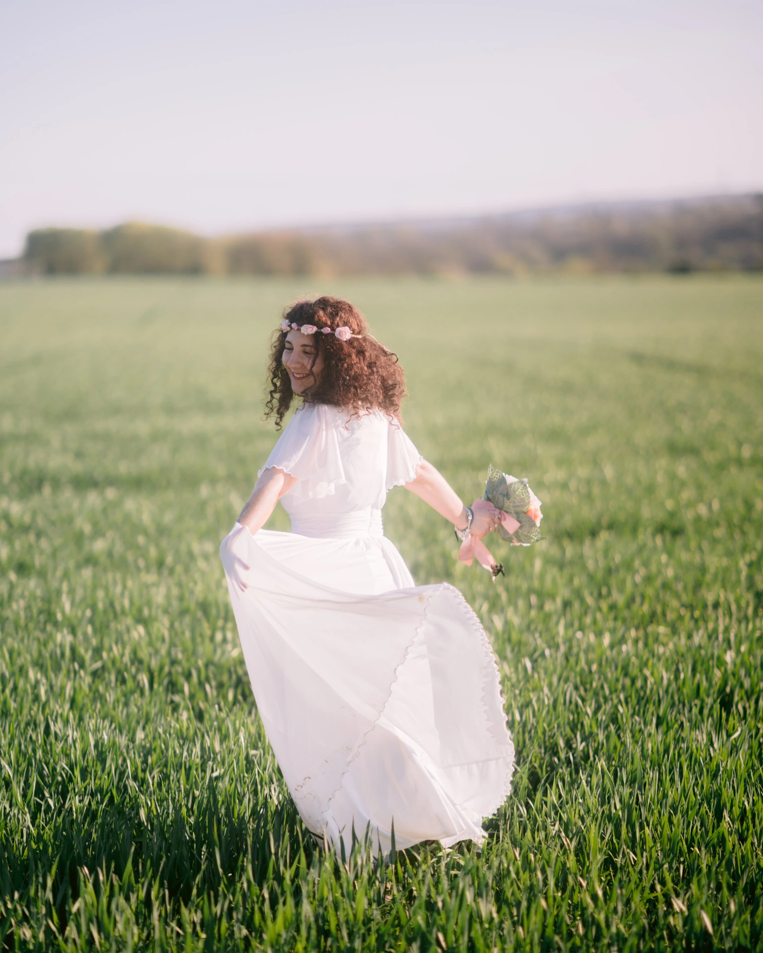 Séance photo lumière naturelle Ruffec Charente - portrait femme dansant dans un champ vert, couronne de fleurs et bouquet, robe blanche qui virevolte au coucher du soleil