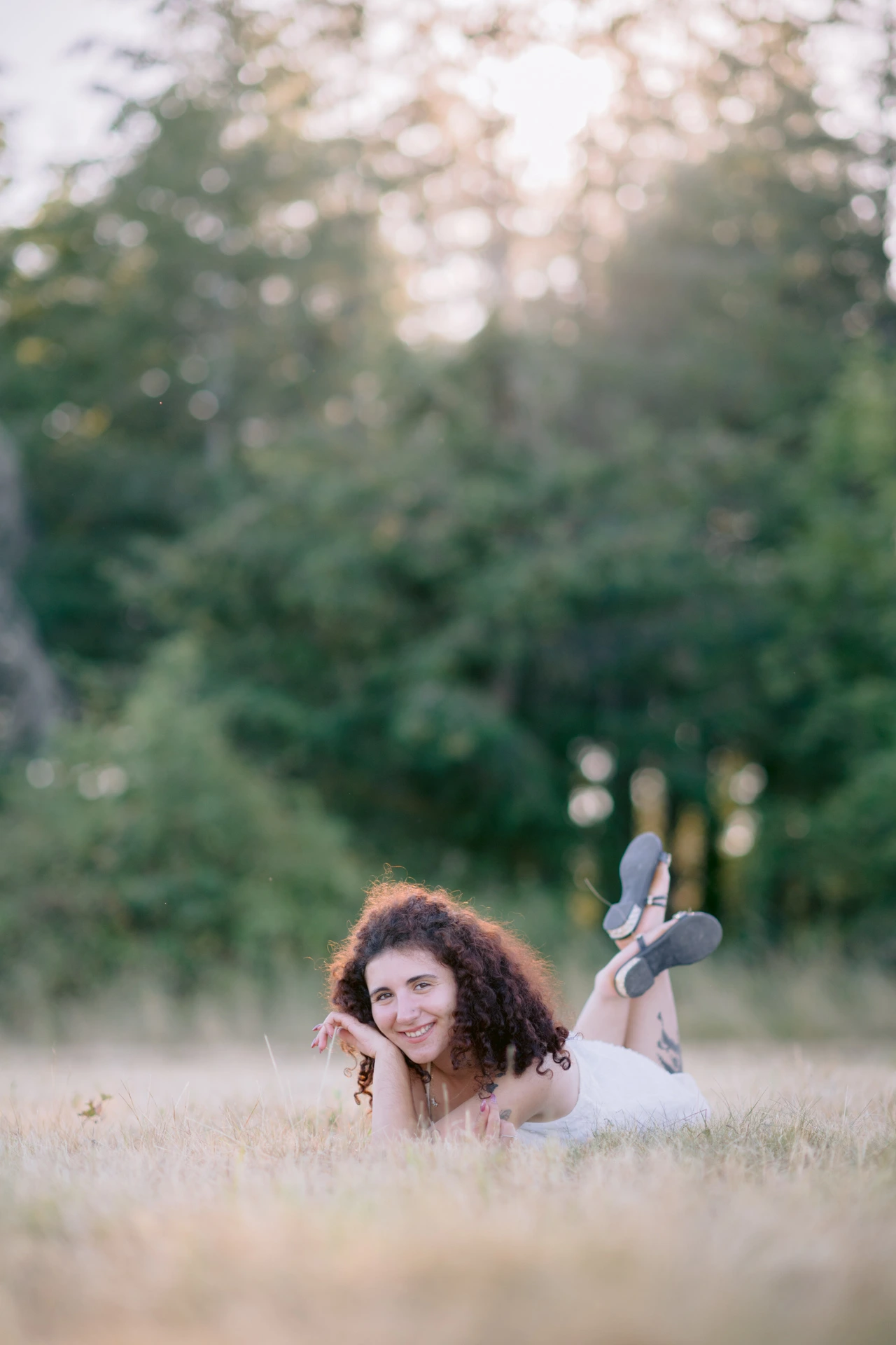 Portrait fine art d'une jeune femme aux cheveux bouclés allongée dans un pré au coucher du soleil, jambes repliées en l'air, à Écuras en Charente