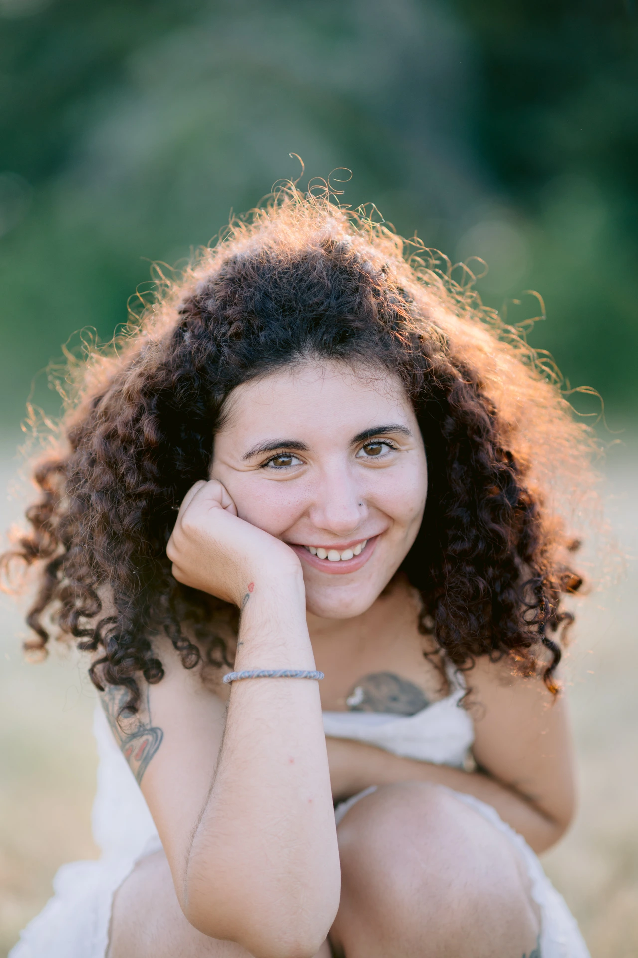 Portrait fine art rapproché d'une jeune femme aux cheveux bouclés souriante, joue posée sur la main, lumière dorée, à Écuras en Charente