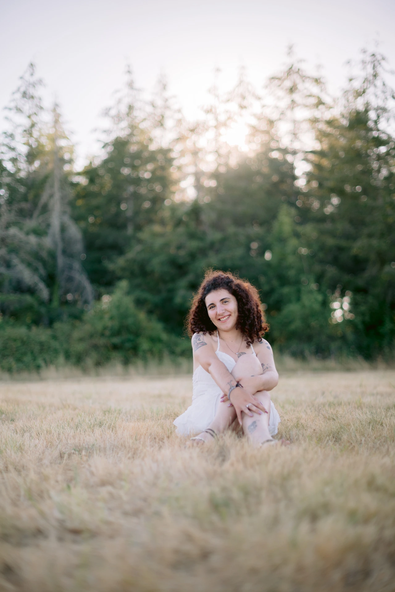 Portrait fine art d'une jeune femme aux cheveux bouclés assise dans un pré au coucher du soleil, robe blanche, à Écuras en Charente